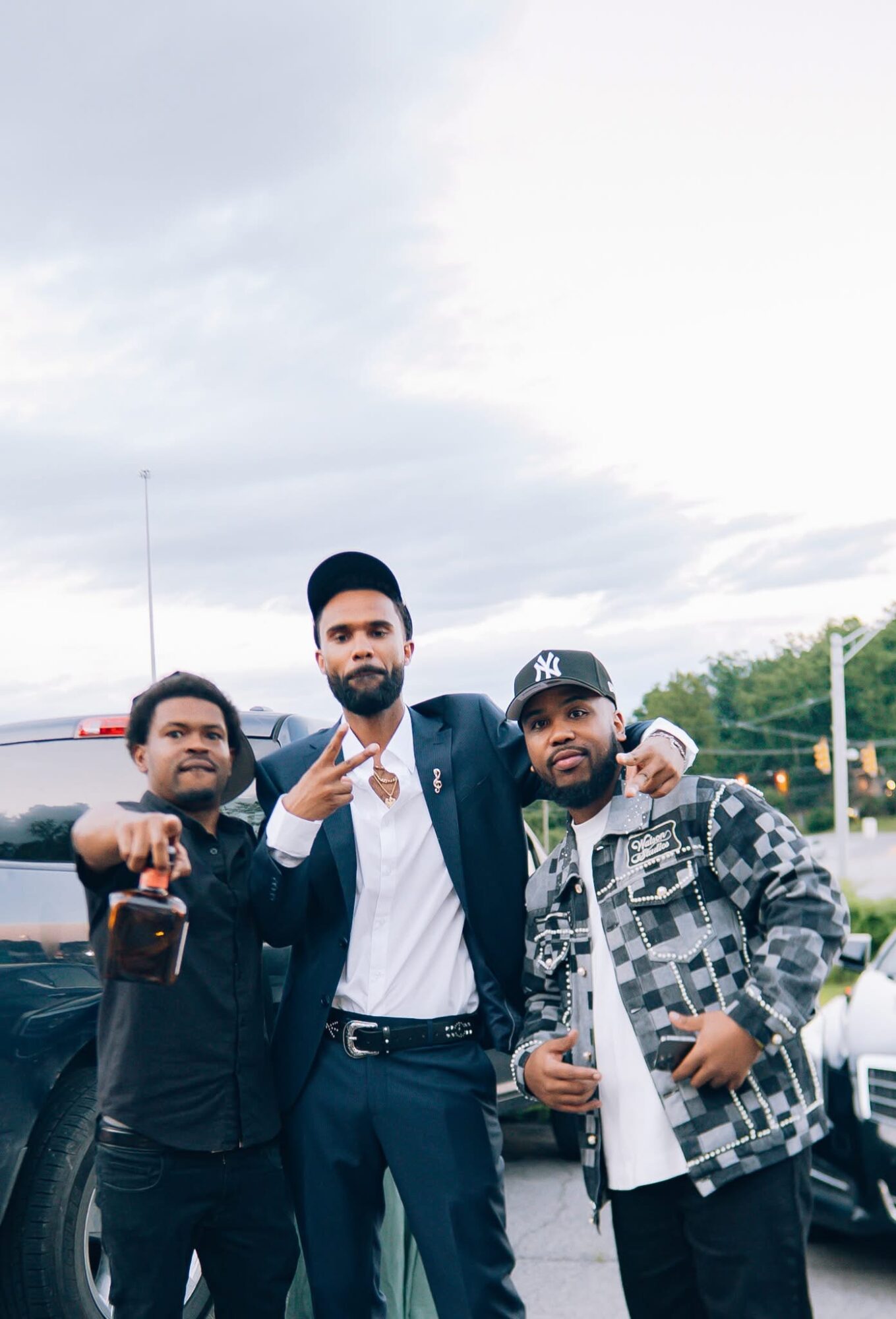Three men standing outdoors, one in a suit, two in casual clothing, with cars and trees in the background.