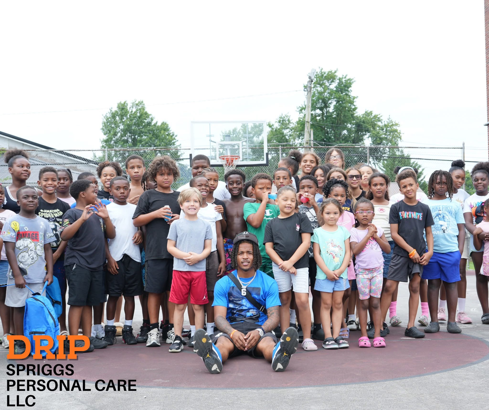 Group of children and adults on a basketball court outdoors, with trees and a fence in the background.