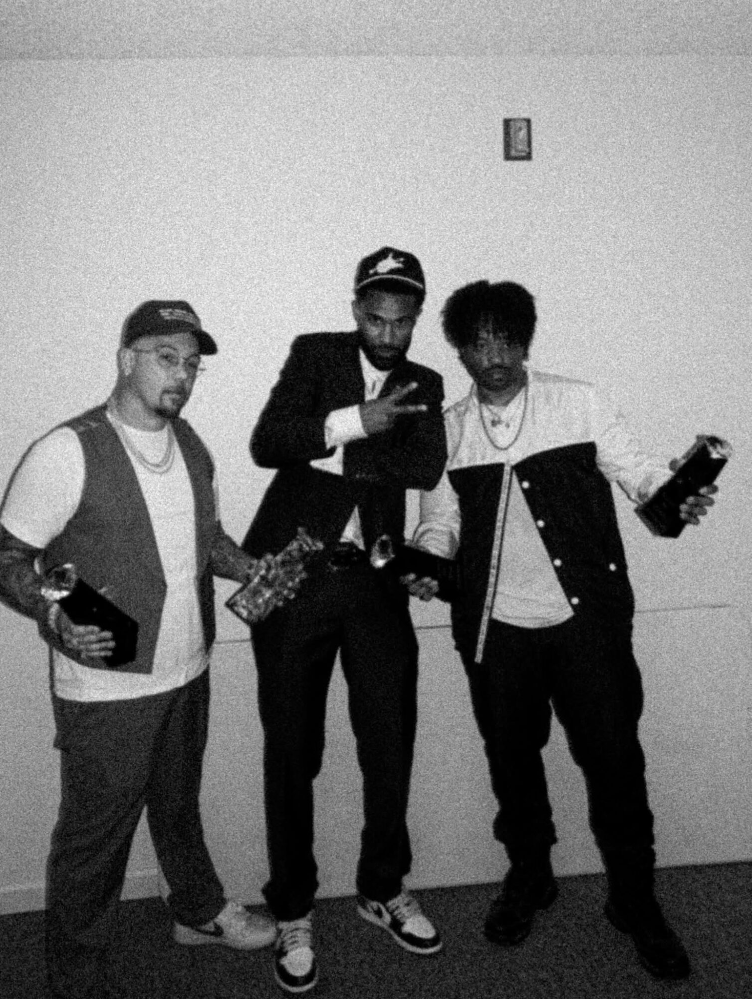 Three people standing against a plain wall, holding objects, with one wearing a cap and glasses, in black and white.