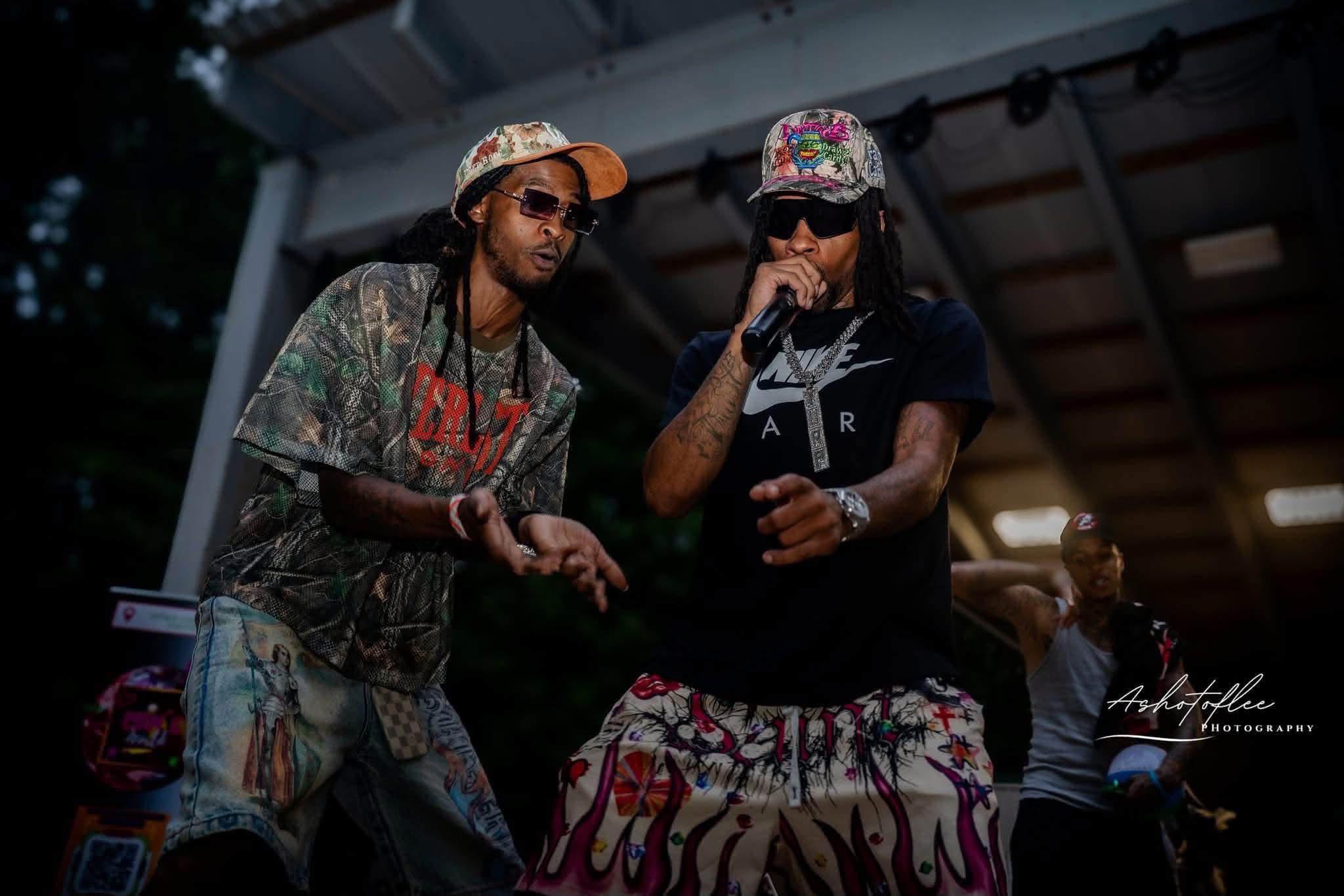 Two men with colorful hats and sunglasses perform on stage under a roof, with a person in the background. Night setting.