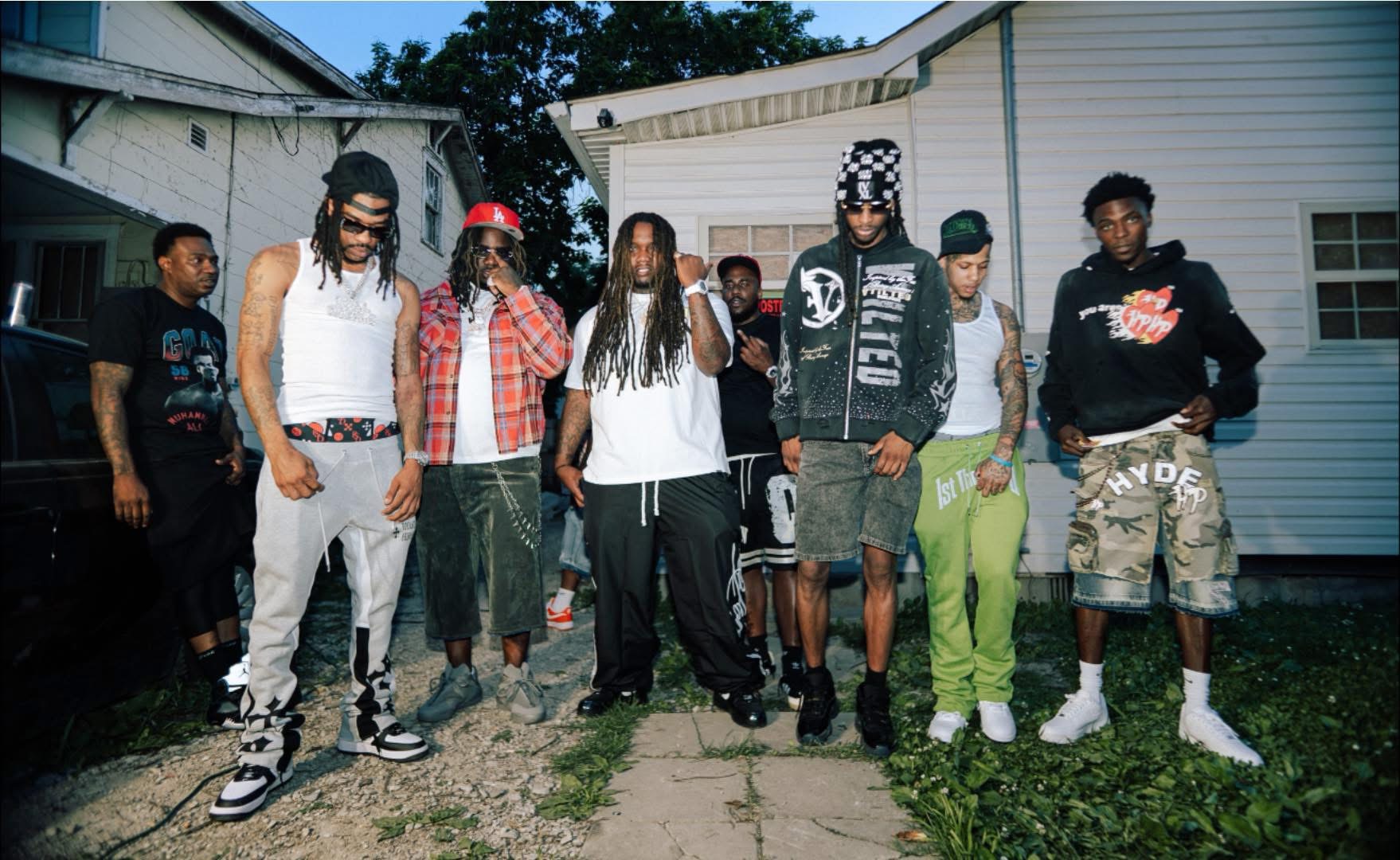 Group of people standing outdoors in front of houses, some wearing casual clothing and accessories, on a sunny day.