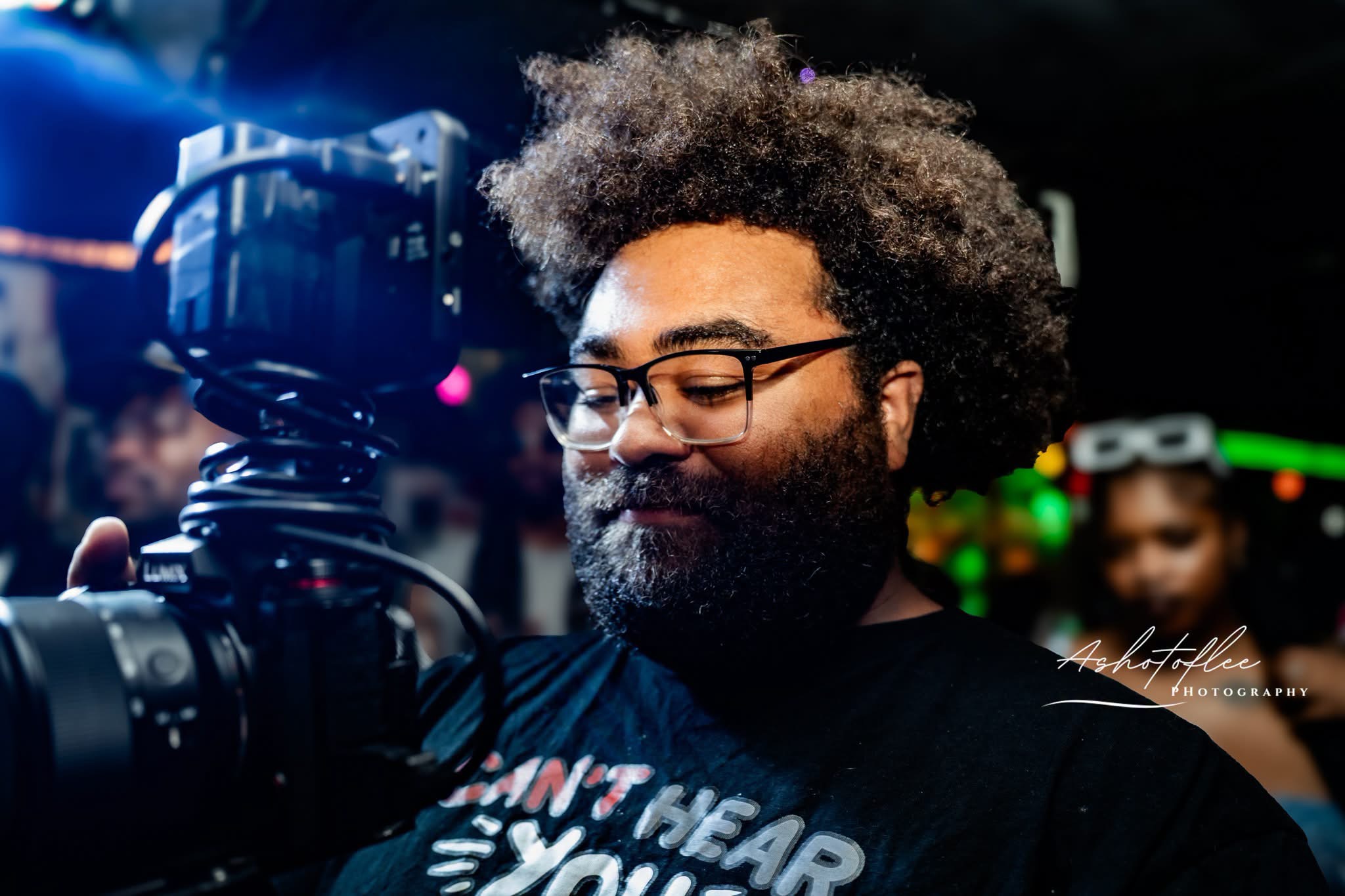 Man with glasses and curly hair operating a camera in a dimly lit environment.