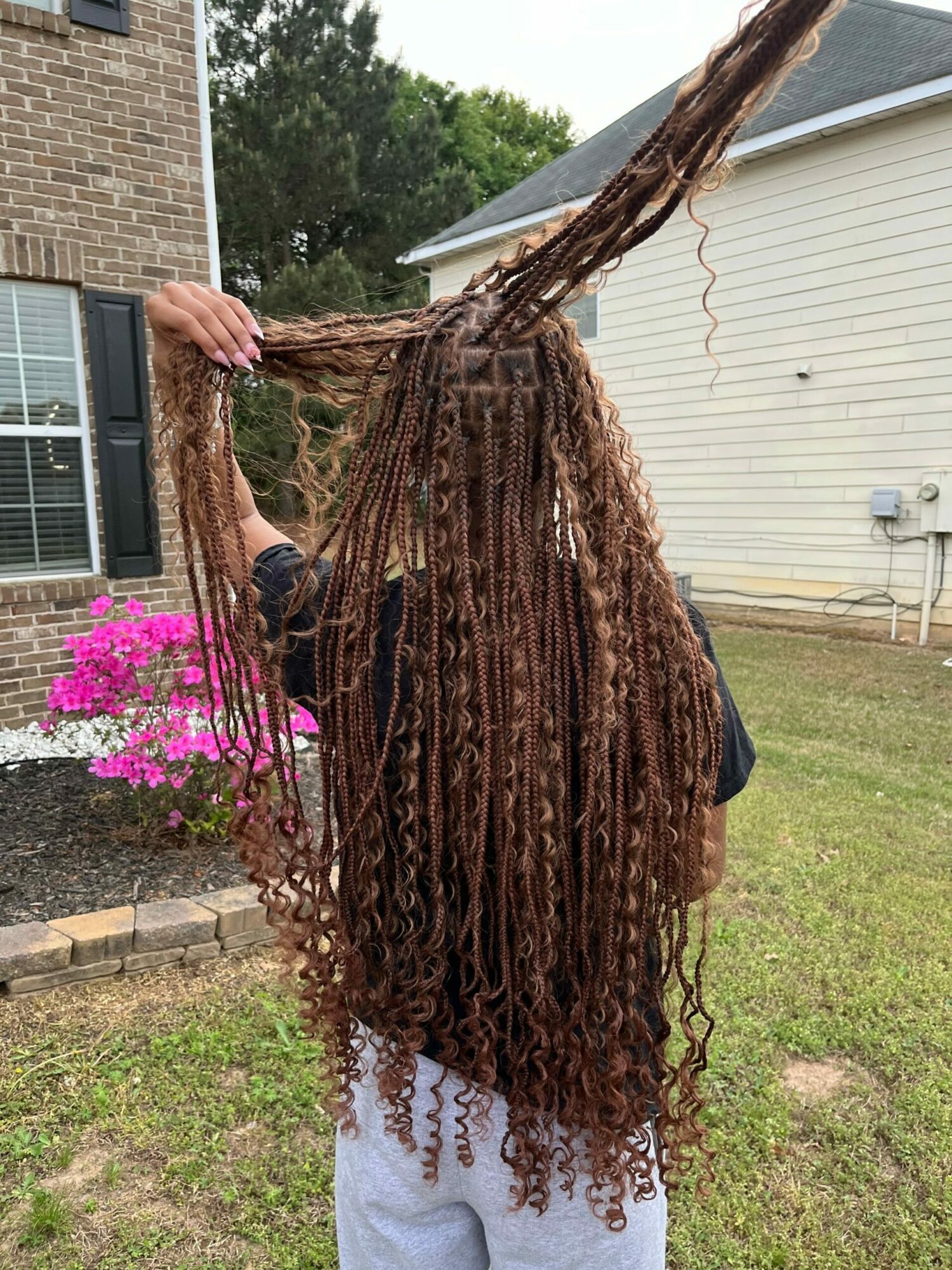 Person with long, curly hair outdoors near a house with pink flowers in the background.