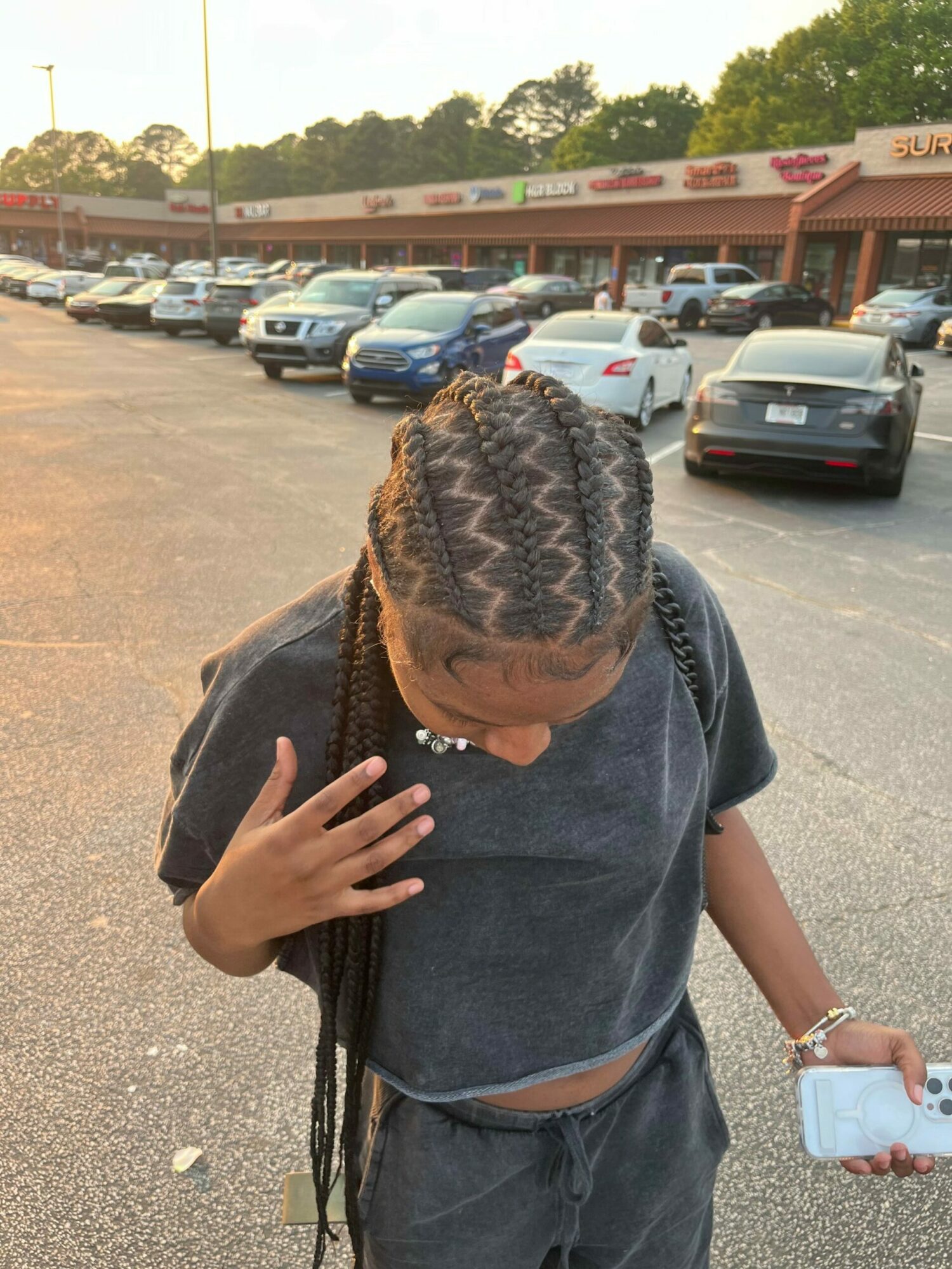 Young person with braided hair walking in parking lot holding phone, wearing dark t-shirt and pants, with cars in background.