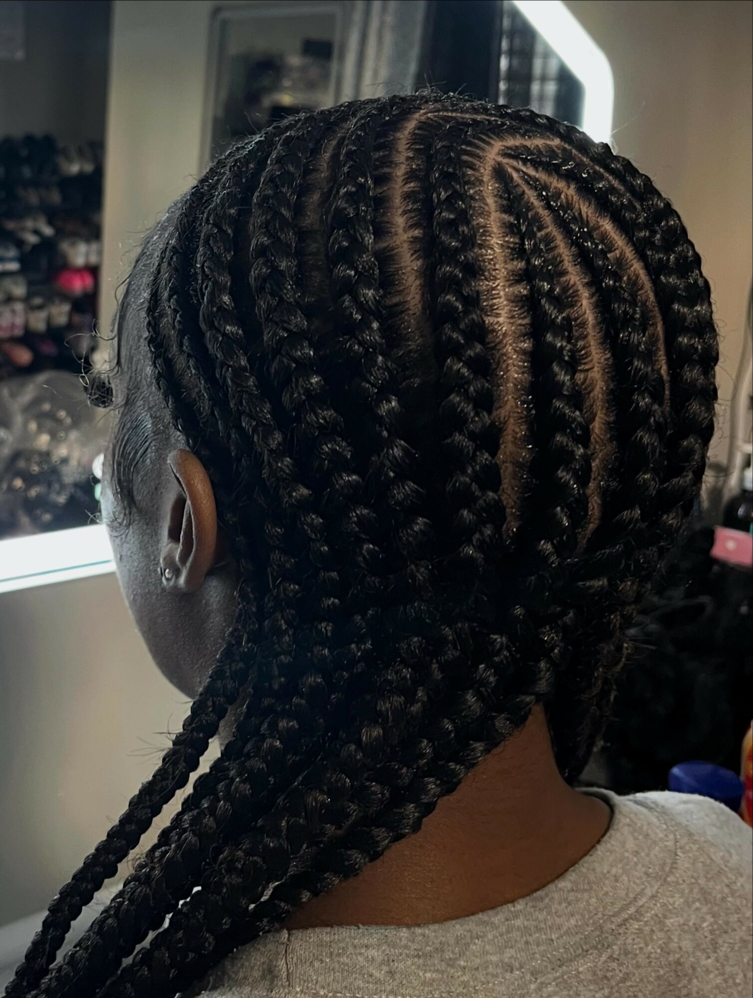 Back of a person's head with braided hairstyle, showing intricate cornrows and long braids, indoors near a mirror.