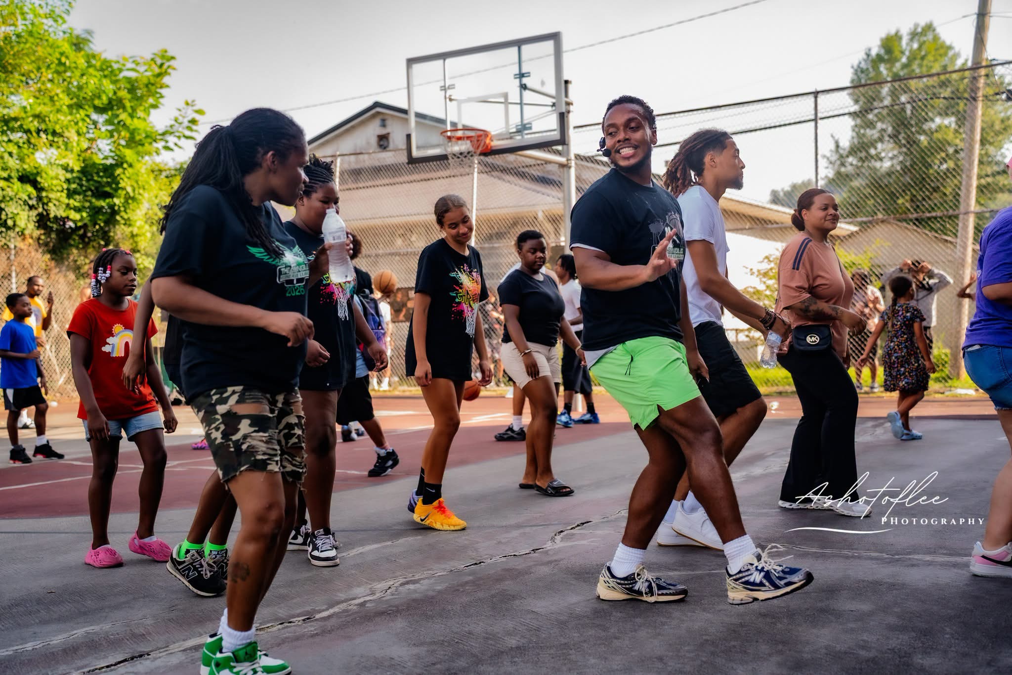 Group of children and adults playing basketball outdoors on a court with a basketball hoop, trees, and buildings in background.