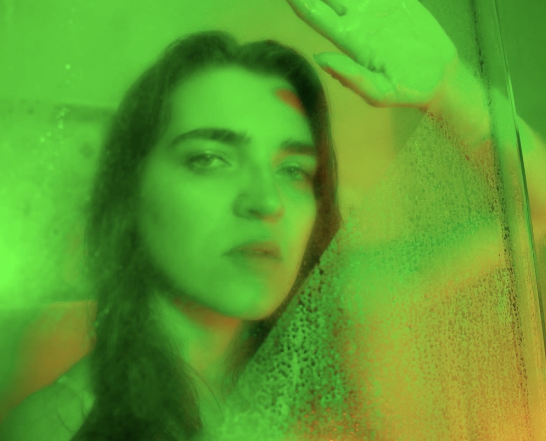 Person with long hair looking at camera, hand raised, behind a green-tinted glass or surface.