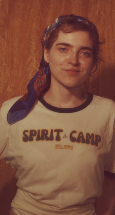Young woman with a headscarf, smiling, wearing a T-shirt with text, standing against a wooden background.