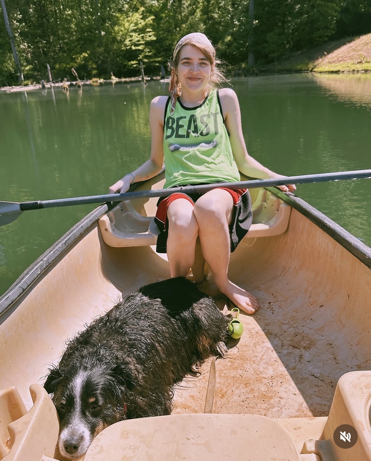 Girl sitting in a canoe with a black and white dog, on a lake with trees in the background.