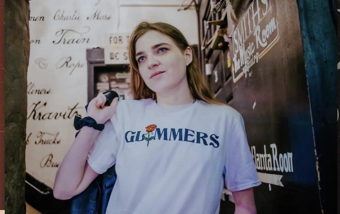 Young woman with brown hair wearing a white T-shirt with blue text, standing indoors near a blackboard and handwritten notes.