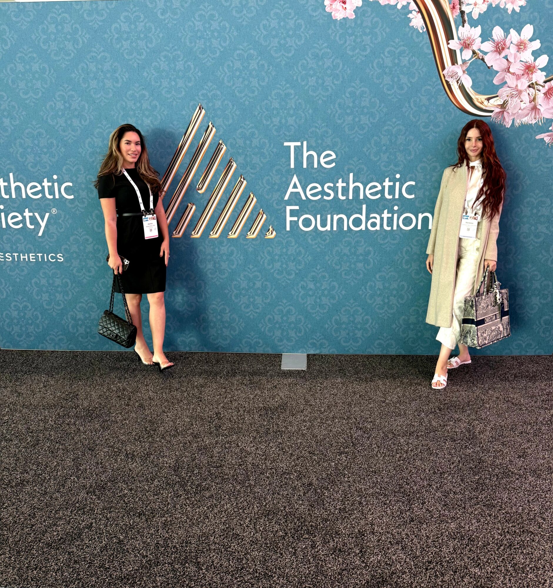 Two women stand in front of a blue wall with text and a logo, holding bags, at an event. Word count: 20.