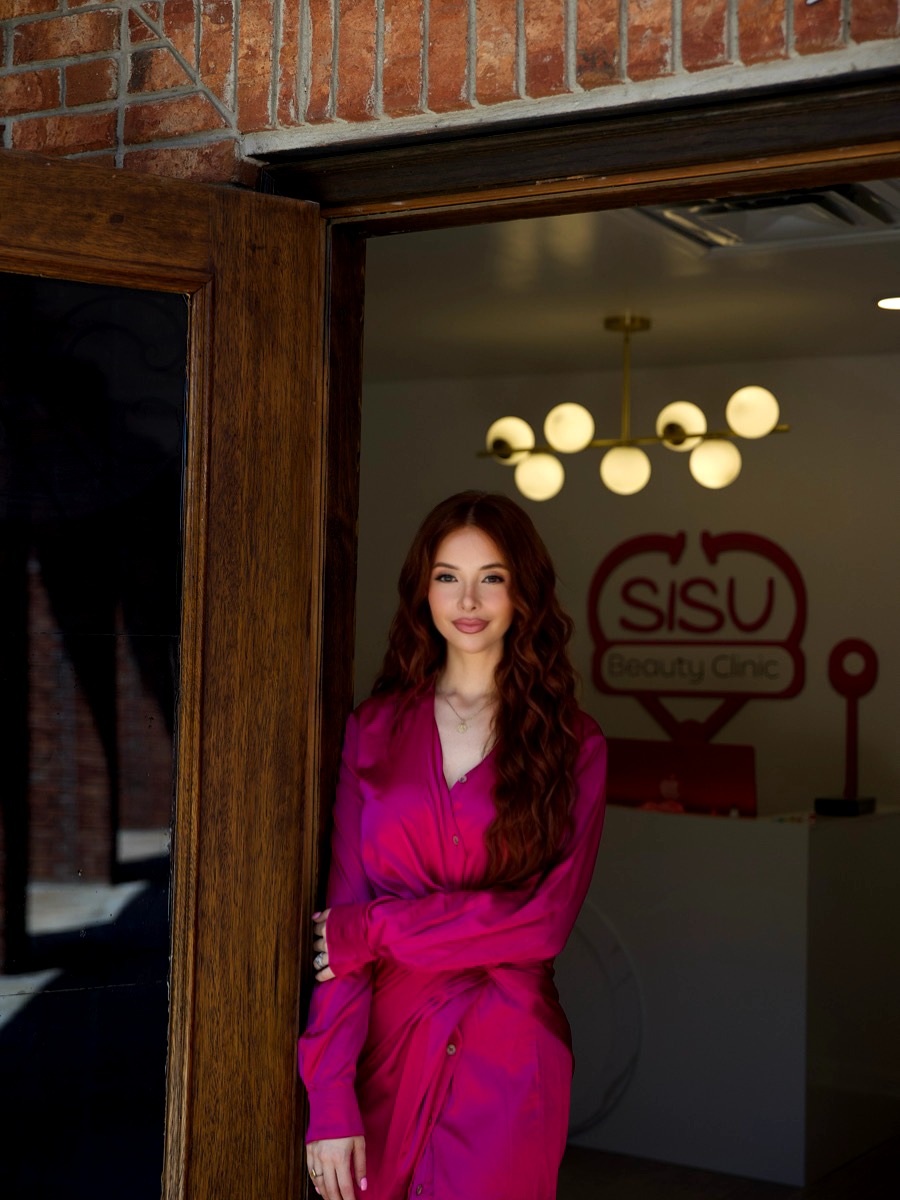 Woman with long curly hair in a pink dress standing inside a building near a brick wall and glass door.