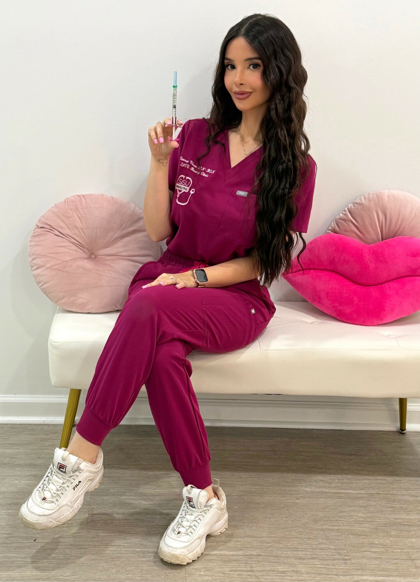 Young woman in pink scrubs sitting on a white bench with pink pillows, holding a syringe, smiling, wearing white sneakers.