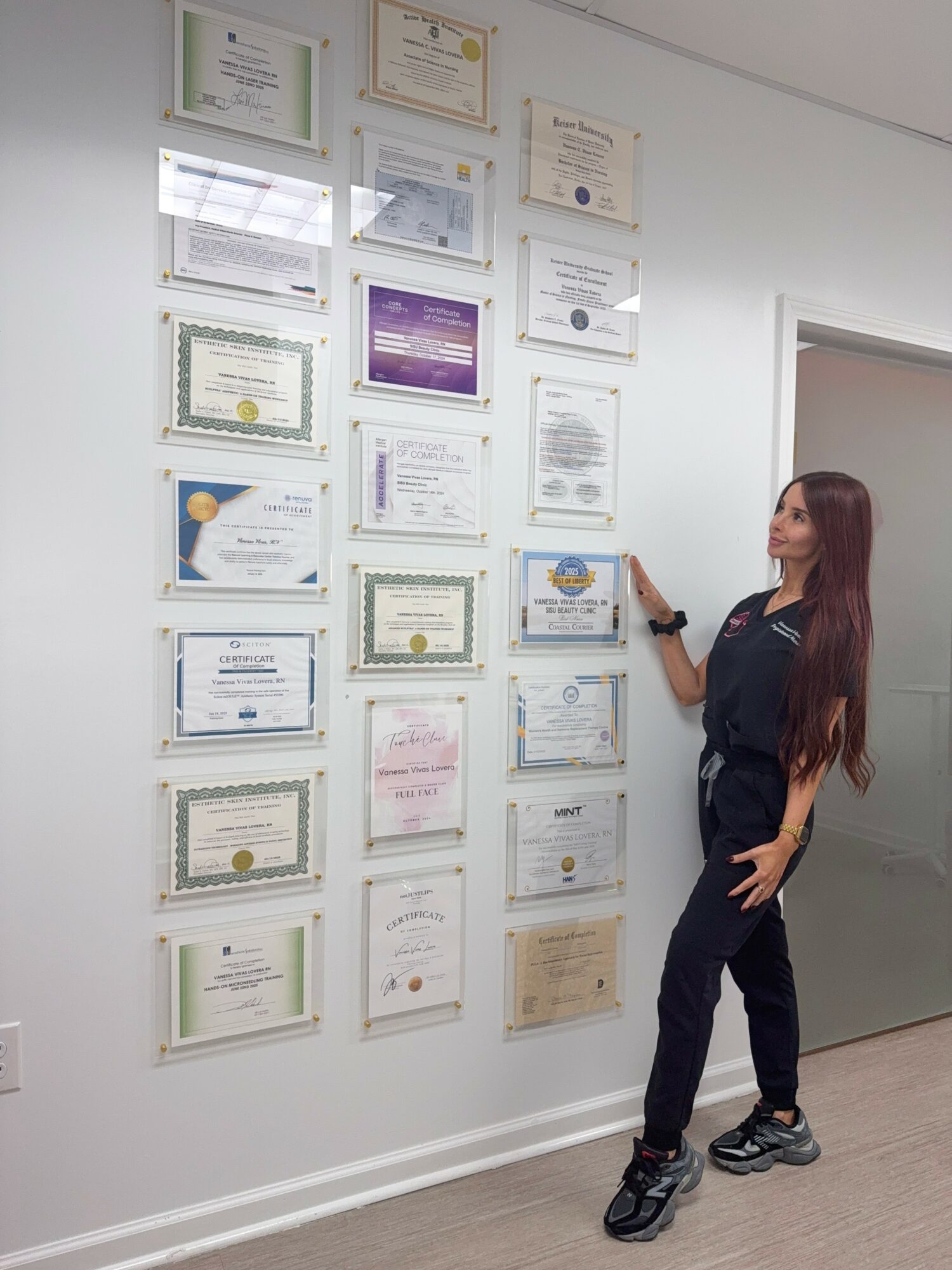 Woman with long hair in dark clothing points at framed certificates on a white wall.