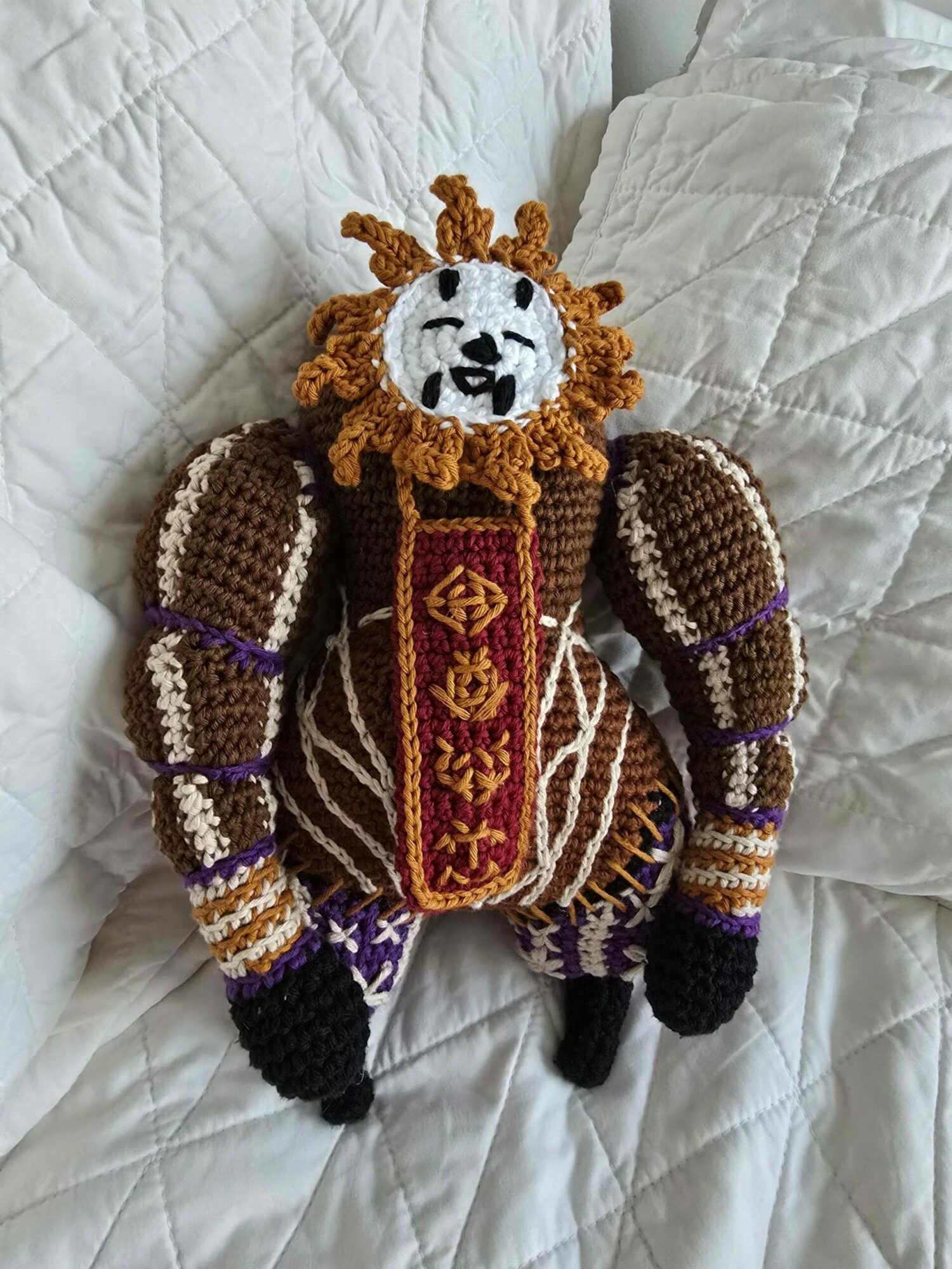 Knitted snake toy with a lion face, lying on a quilted white surface, with detailed patterns and colors.