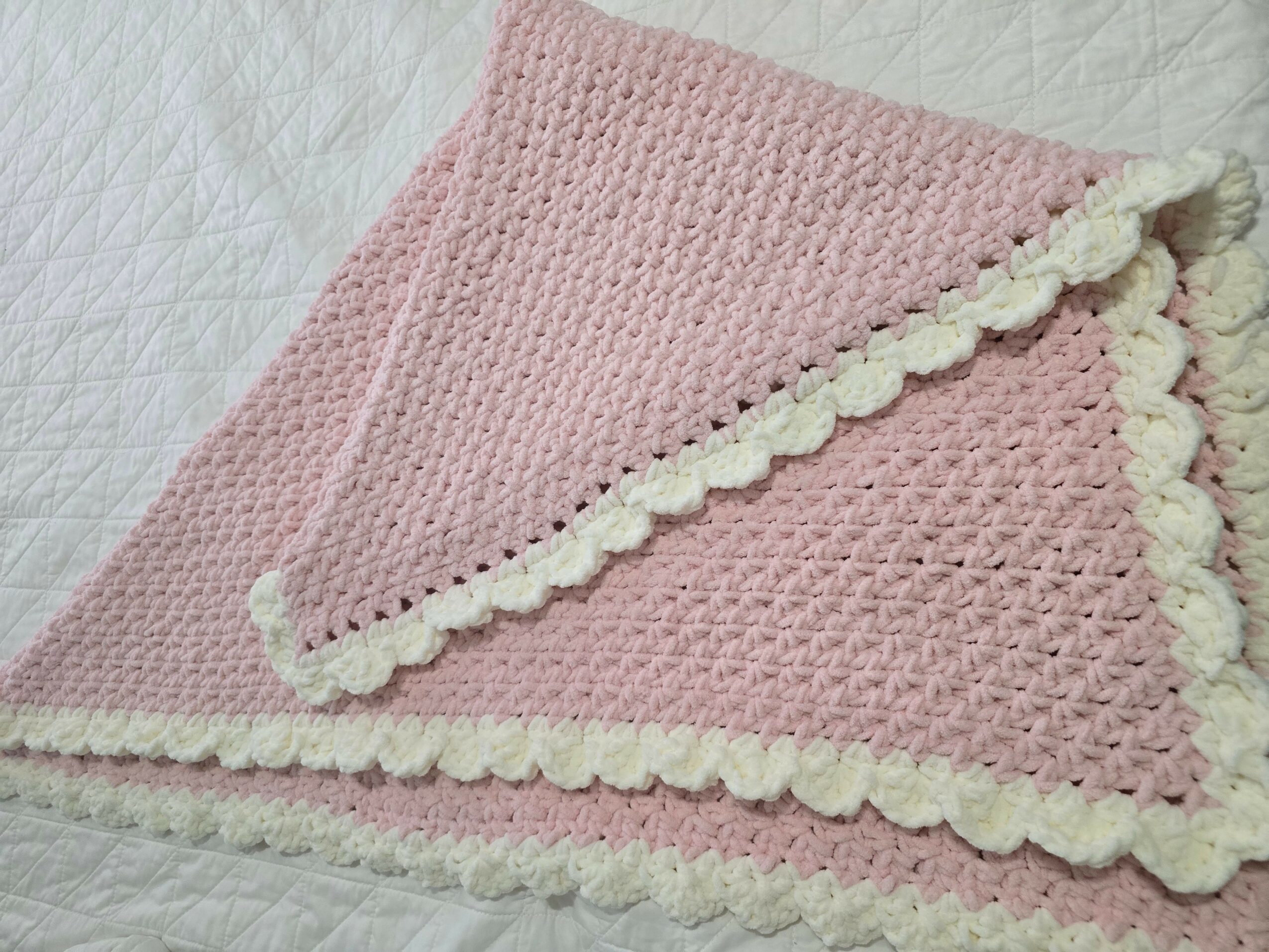 Pink crocheted blanket with white pom-pom border, folded on a white surface.