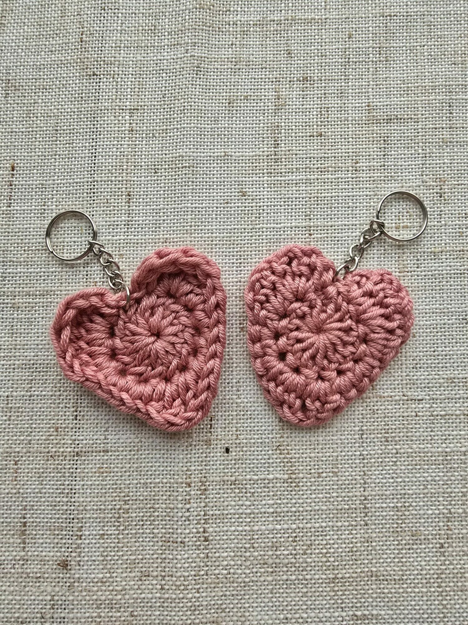 Two pink crocheted heart-shaped keychains on a fabric background, one above the other, with metal key rings attached.