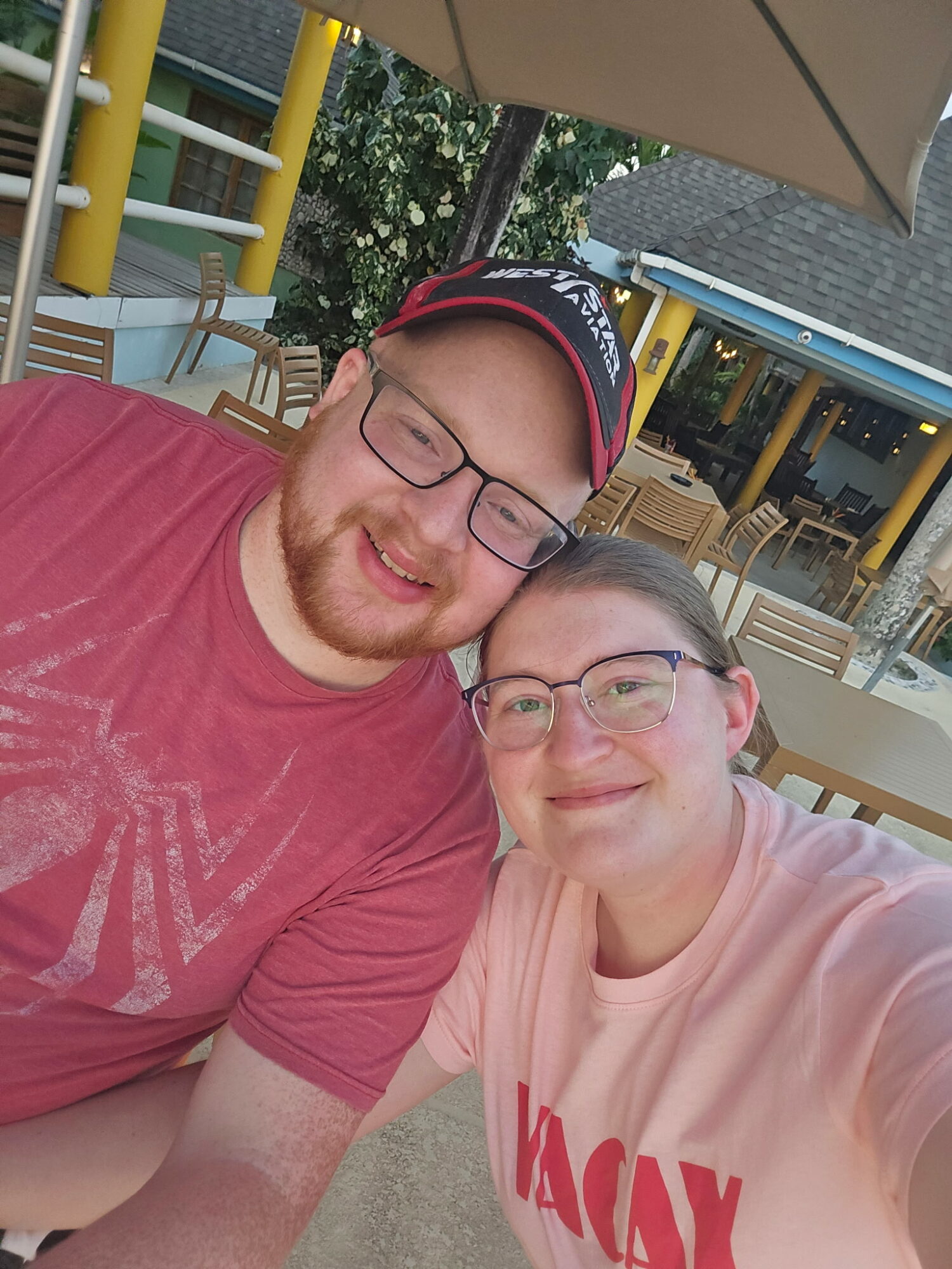 A smiling man and woman wearing glasses take a selfie outdoors at a restaurant patio with tables and chairs, under an umbrella.