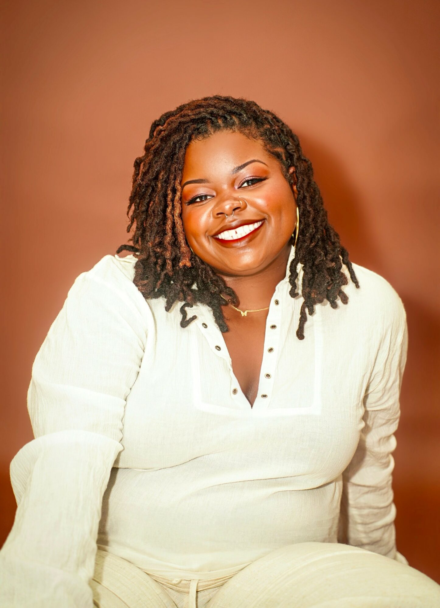 Smiling woman with shoulder-length dreadlocks wearing a white top against a brown background.