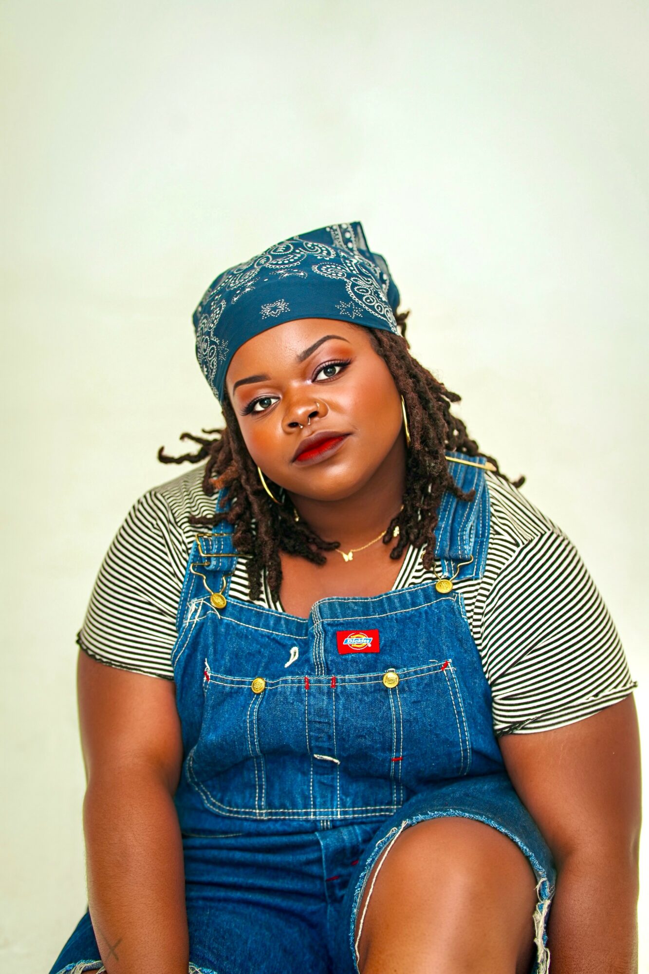 Woman with a headscarf and dreadlocks wearing a striped shirt and denim overalls, looking at the camera.