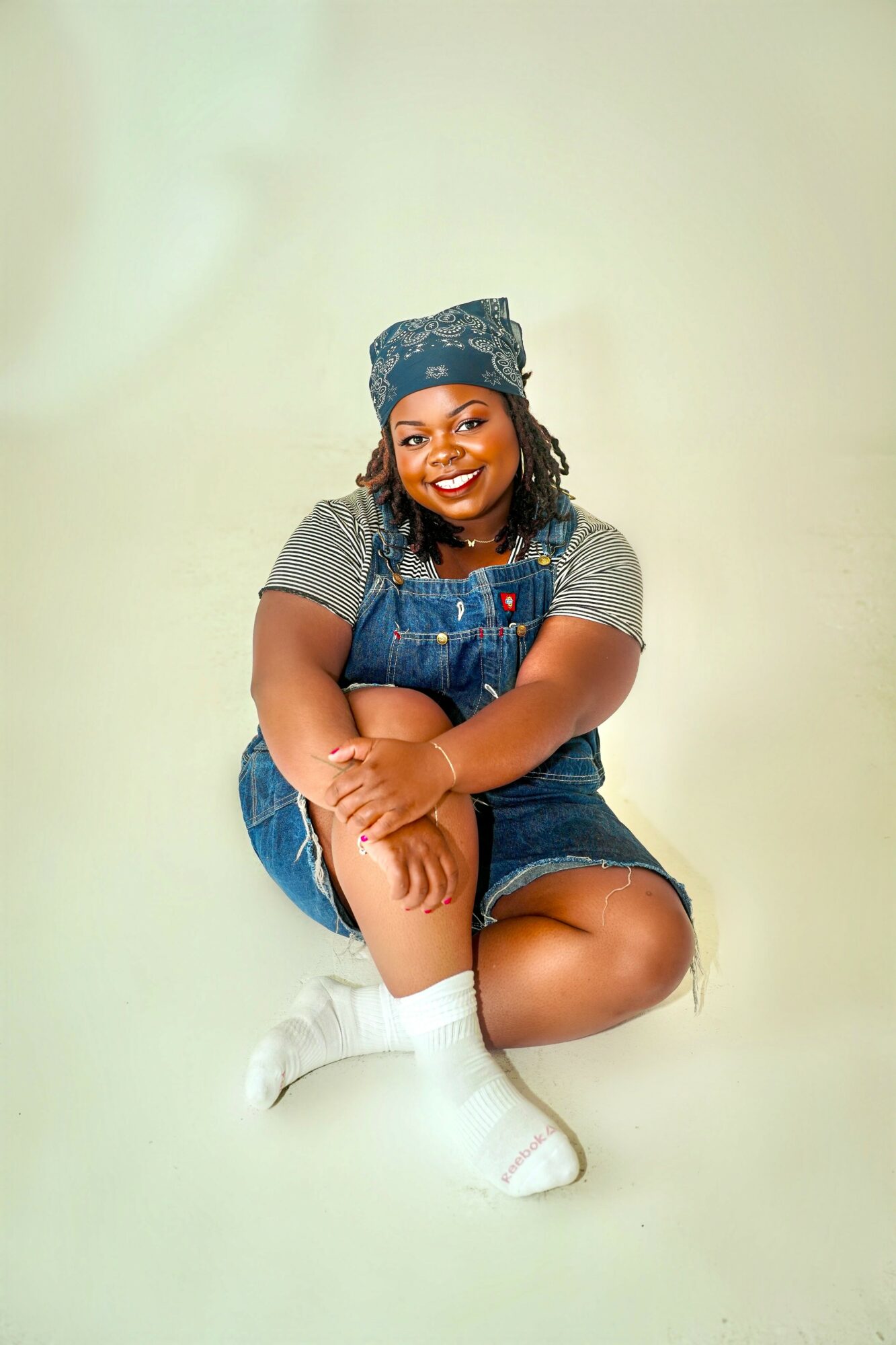 Smiling woman sitting with crossed legs, wearing a headscarf, striped shirt, denim overalls, and white socks, against a light background.