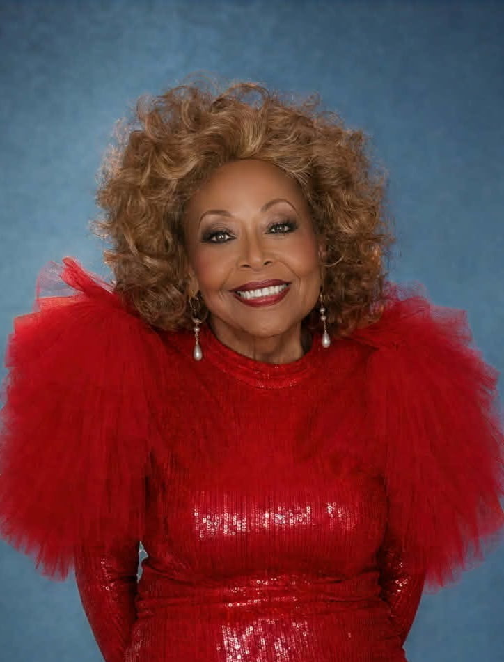 Woman with curly hair wearing a red dress with feathered shoulders, smiling, against a blue background.