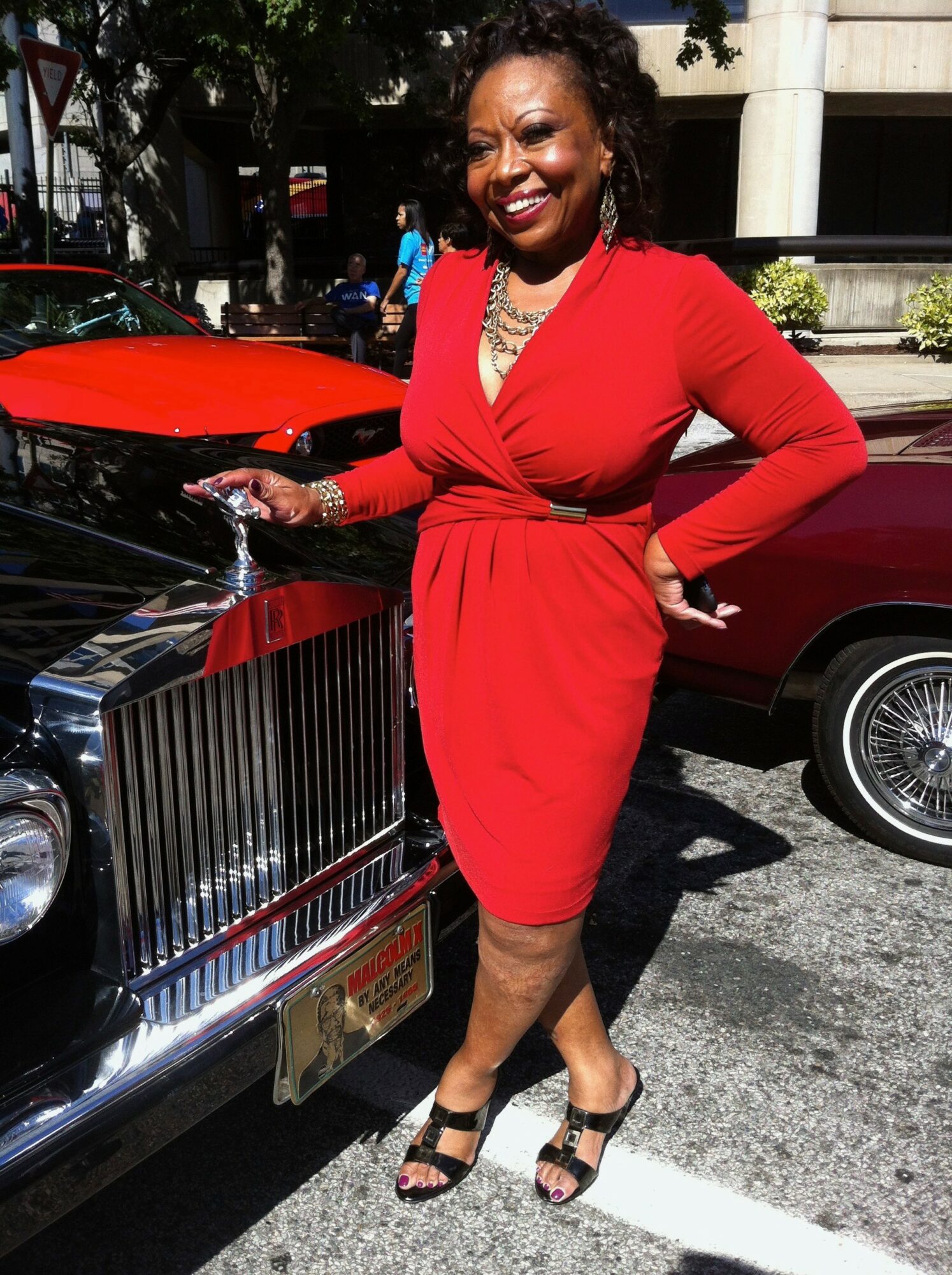 Woman in red dress standing next to a vintage car, smiling, with cars and trees in background.
