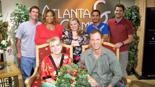 Group of six people in a TV studio, some sitting and some standing, with a floral arrangement in front.