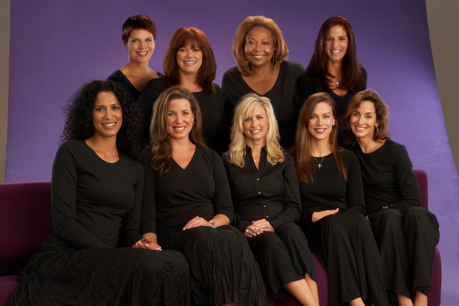 Group of nine women sitting and standing on a purple background, all wearing black outfits, smiling.