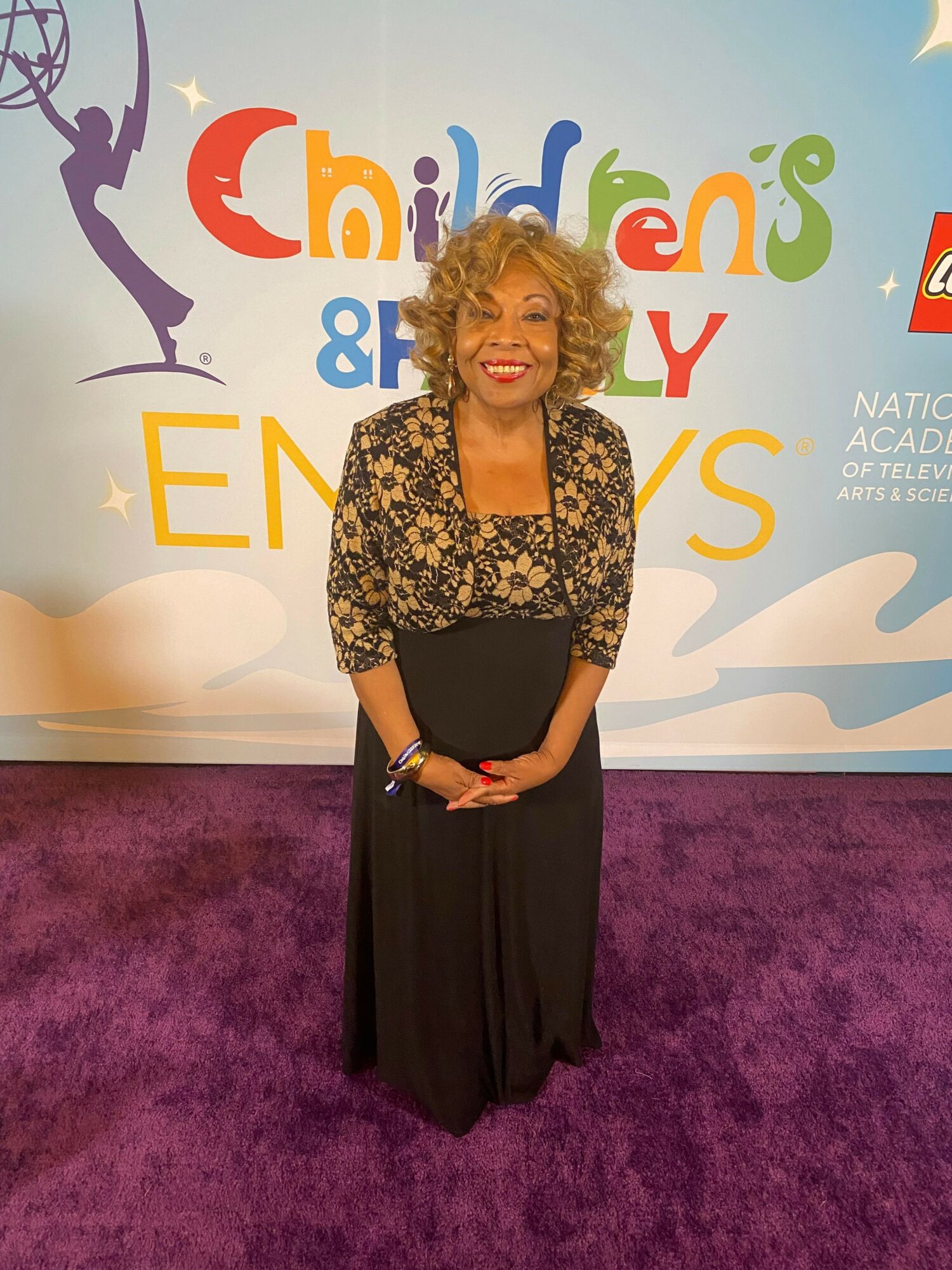 Woman with curly hair in a floral top and black skirt standing on purple carpet, smiling, in front of a colorful backdrop.
