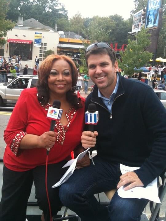 Two people smiling, holding microphones, outdoors in a busy urban area with trees and buildings in background.