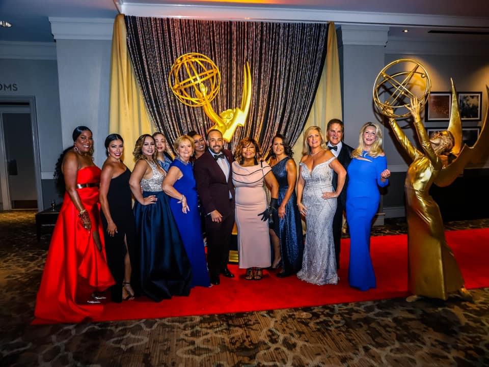 Group of people in formal attire posing in front of a curtain with Emmy awards statues, on a red carpet.