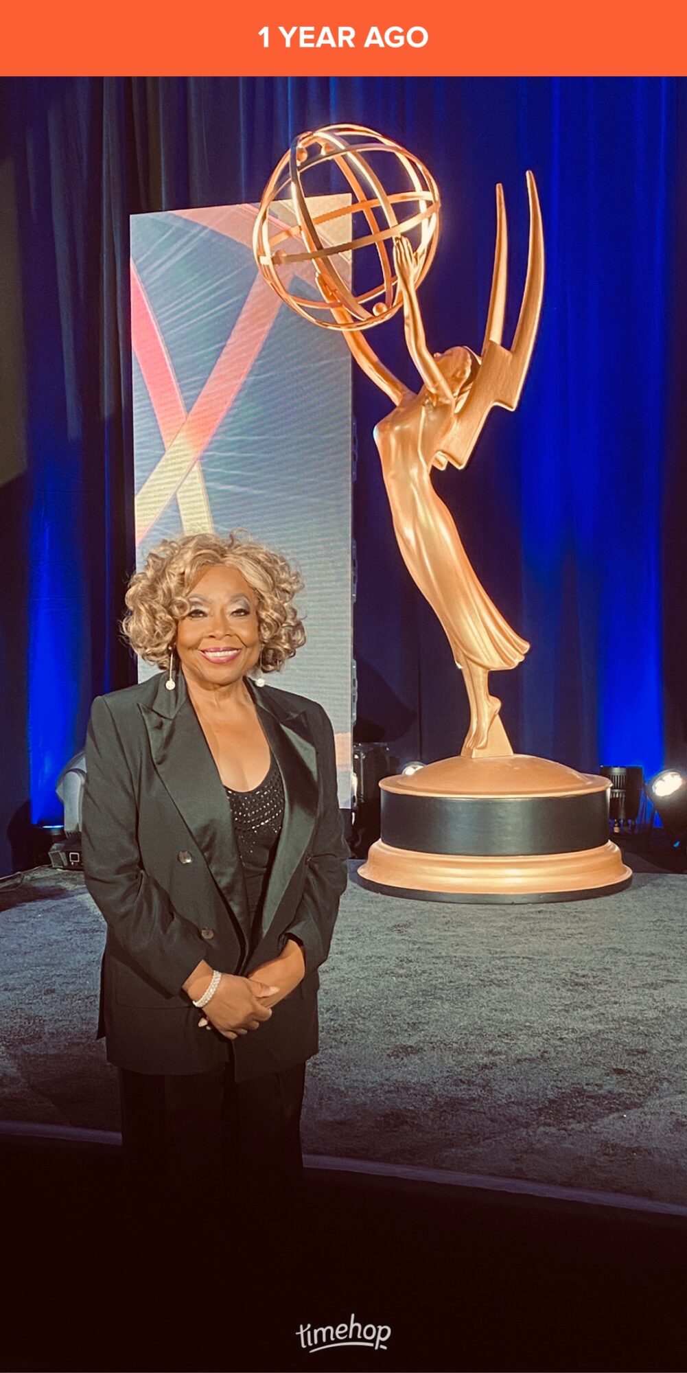 Woman with curly hair in a black blazer standing in front of a large Emmy award statue on stage.