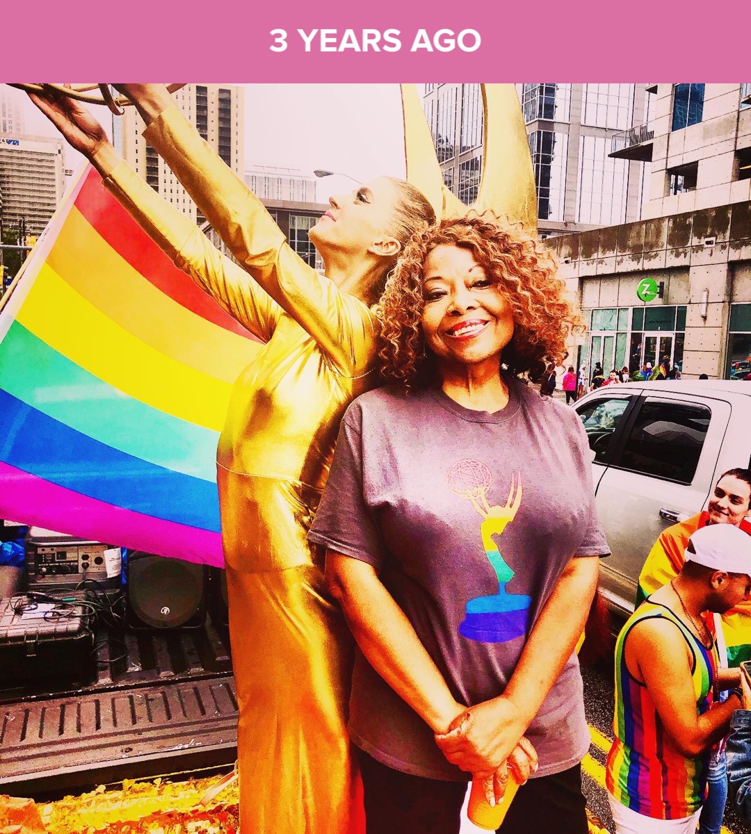 Two women smiling, one holding a rainbow flag, in an urban setting with buildings and cars, celebrating pride.