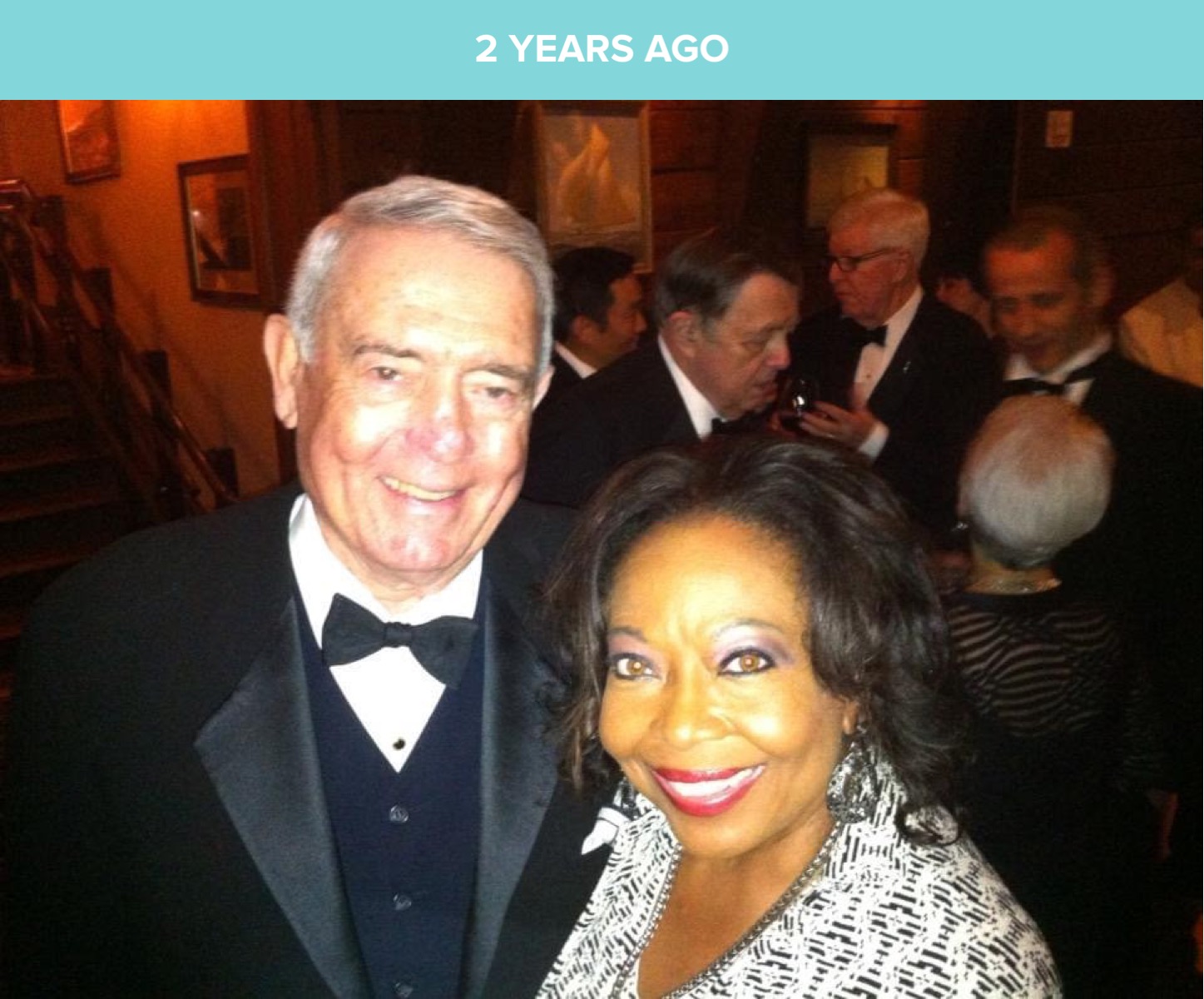 Two people smiling at a formal event, with a group of people in the background, in a warmly lit room.