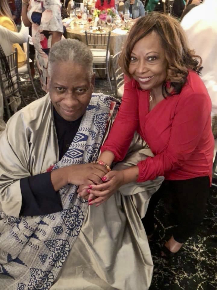 Two women holding hands and smiling at a social event, with tables and people in the background.