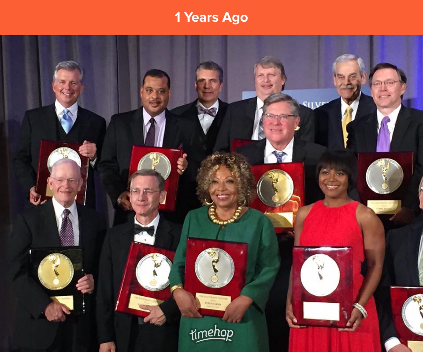 Group of people holding awards, standing on stage with a gray curtain background, some smiling, dressed in formal attire.