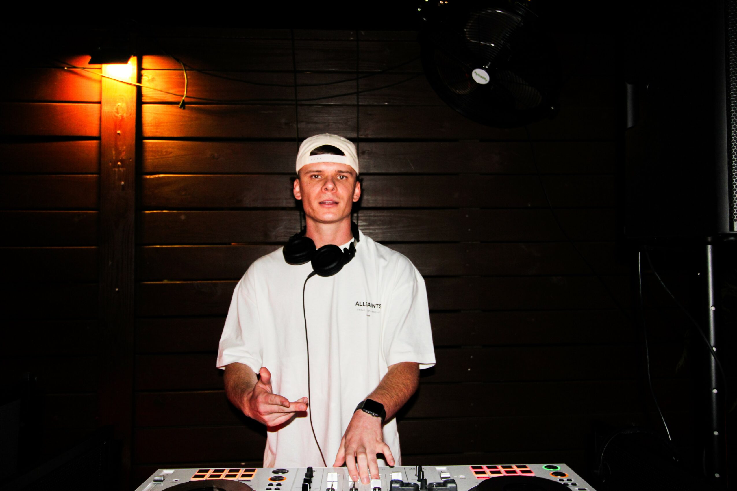 Young man wearing a white shirt, white cap, and headphones around neck, standing behind a DJ mixer in a dark room.