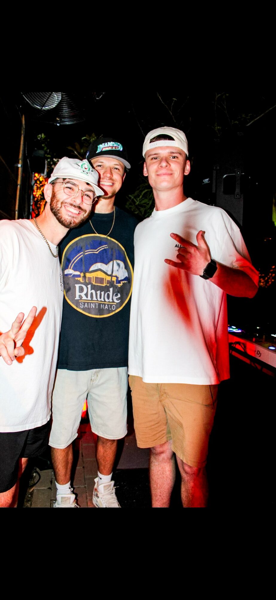 Three young men standing together outdoors at night, smiling, with one making a peace sign, wearing casual clothing and hats.