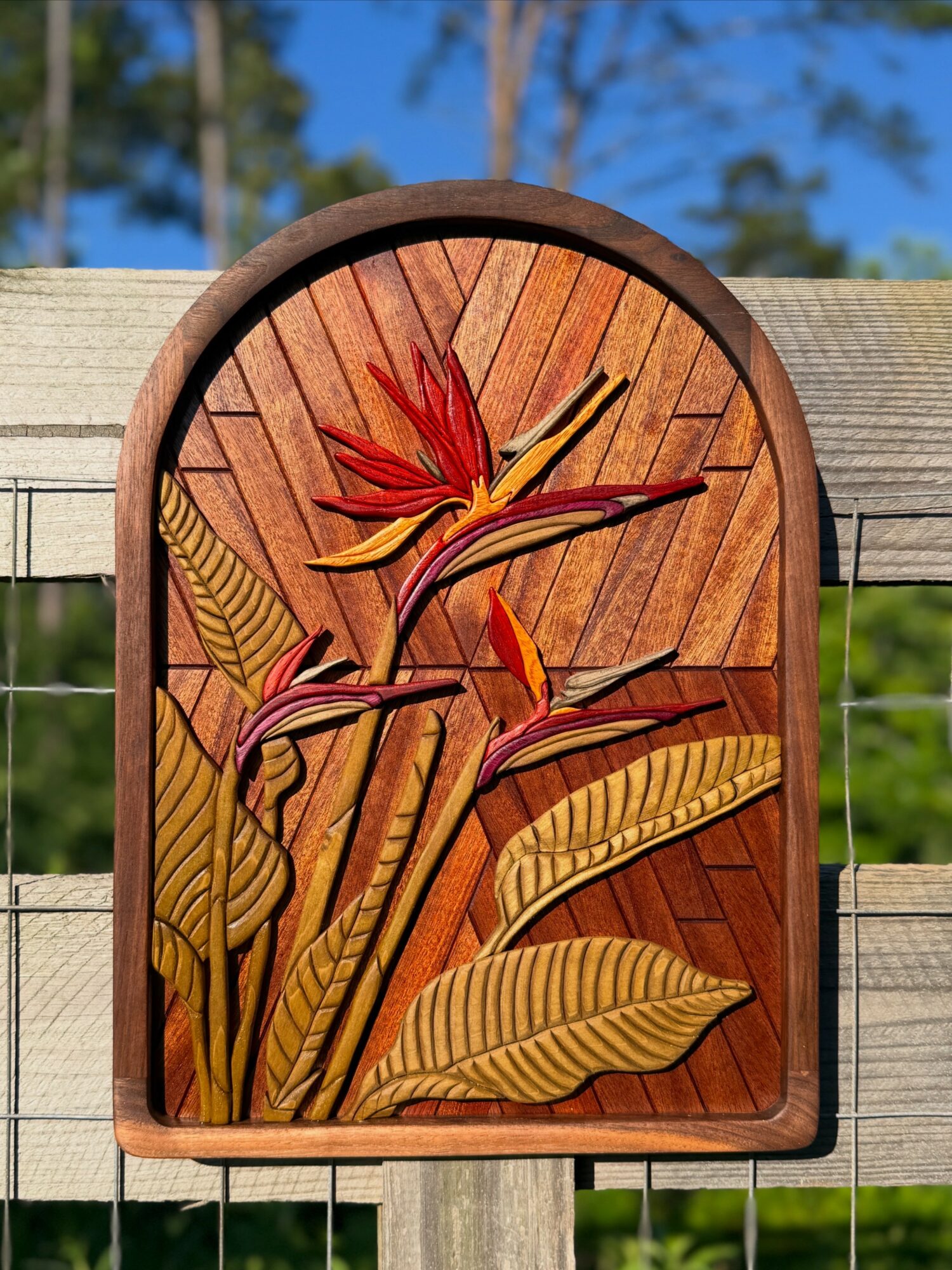 Decorative wooden clock with floral design, featuring large leaves and a flower, mounted on a wooden fence outdoors.