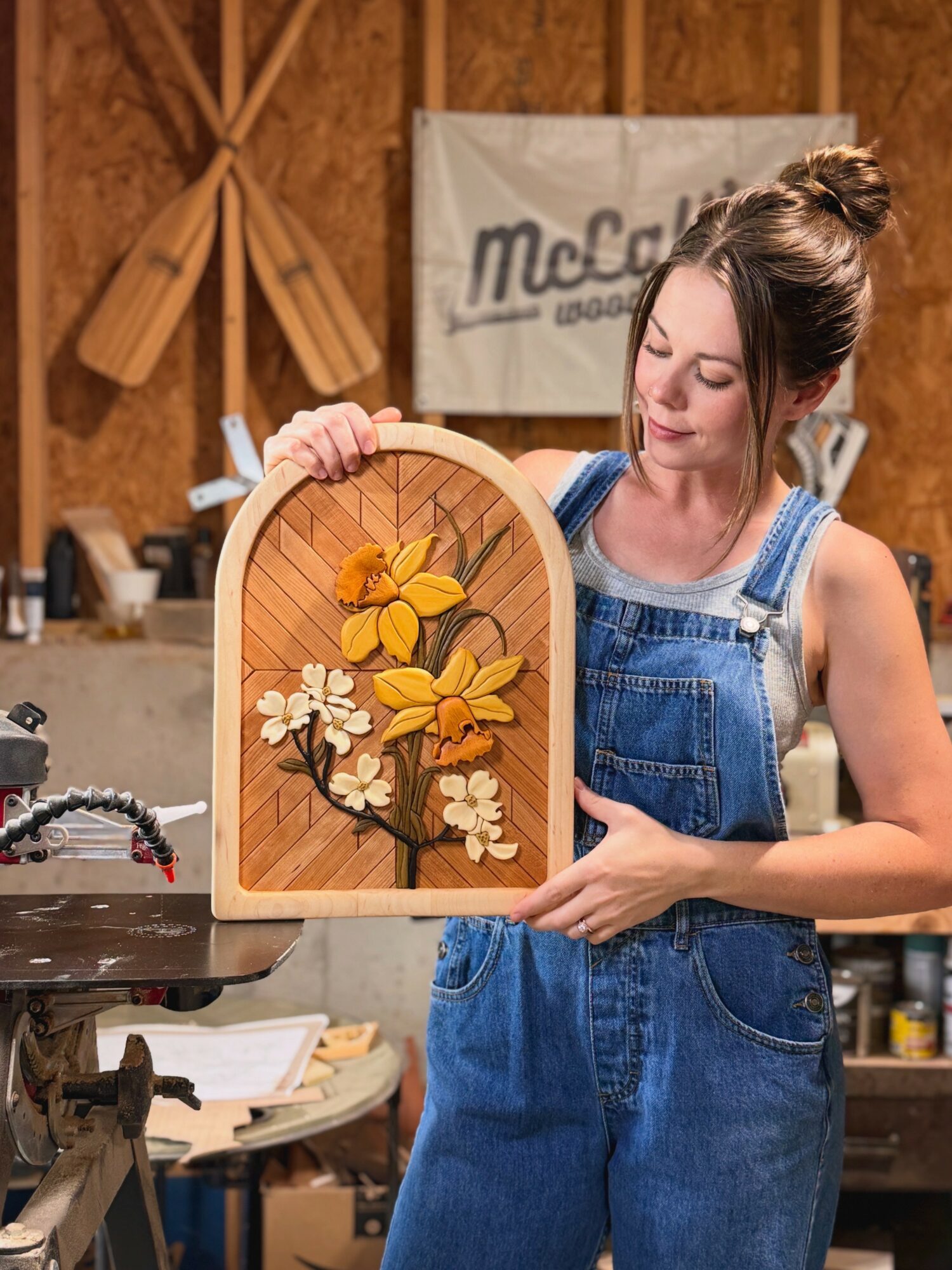 Young woman in denim overalls holds a wooden framed floral artwork in a workshop.