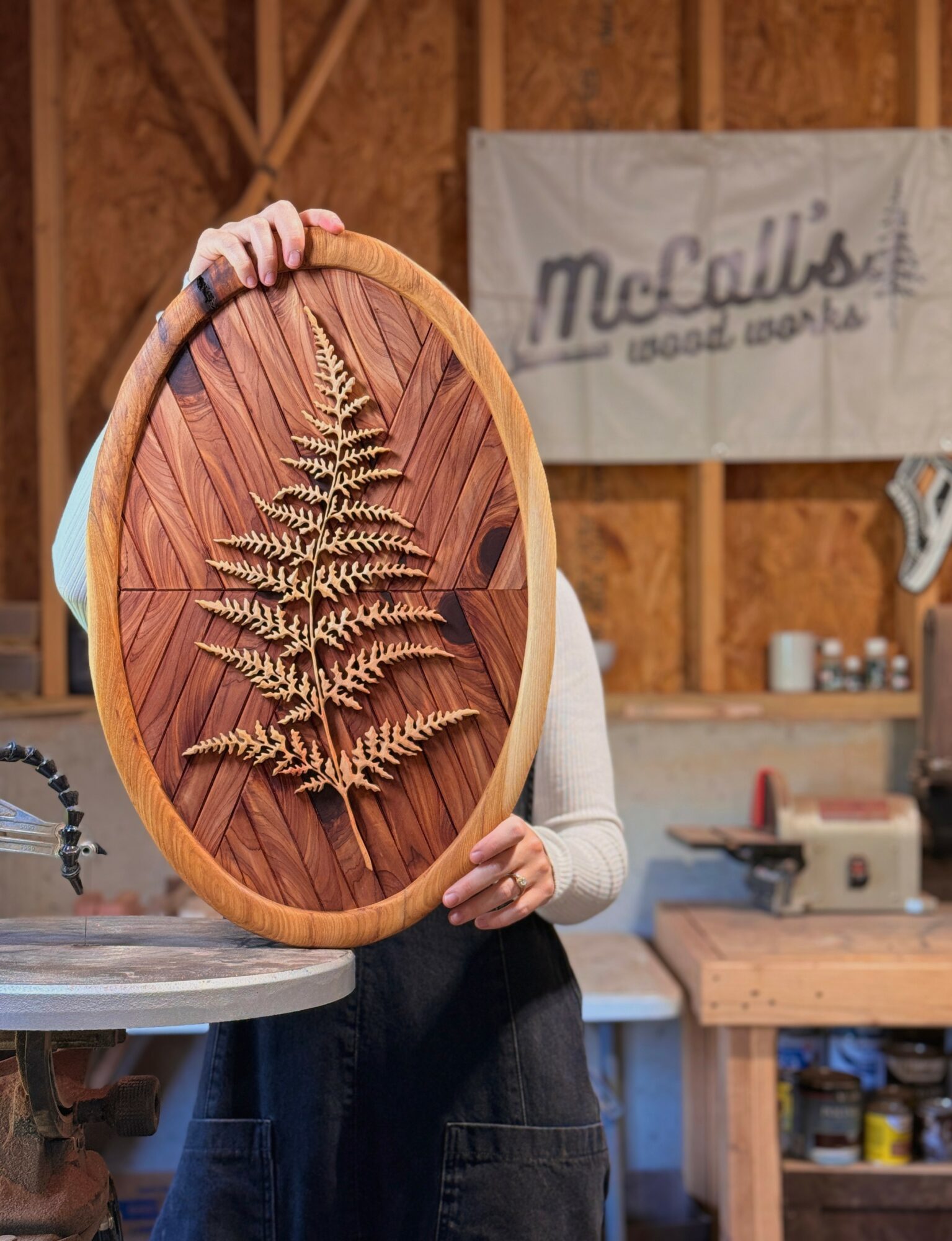 Person holding an oval wooden frame with a fern leaf and wooden slats inside, workshop background with tools and a sign.