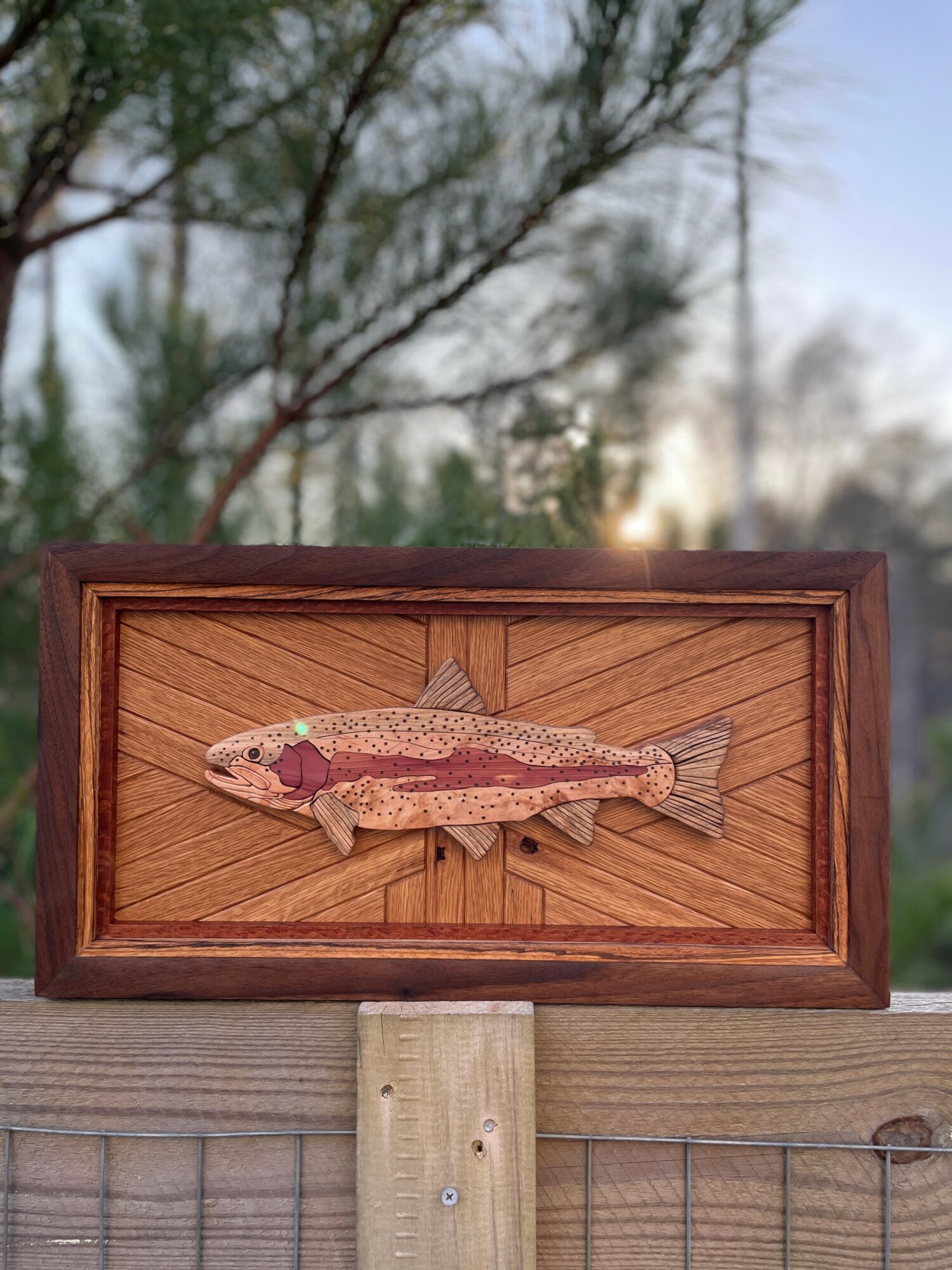 Framed fish artwork mounted on a wooden post outdoors, with trees and sky in background.
