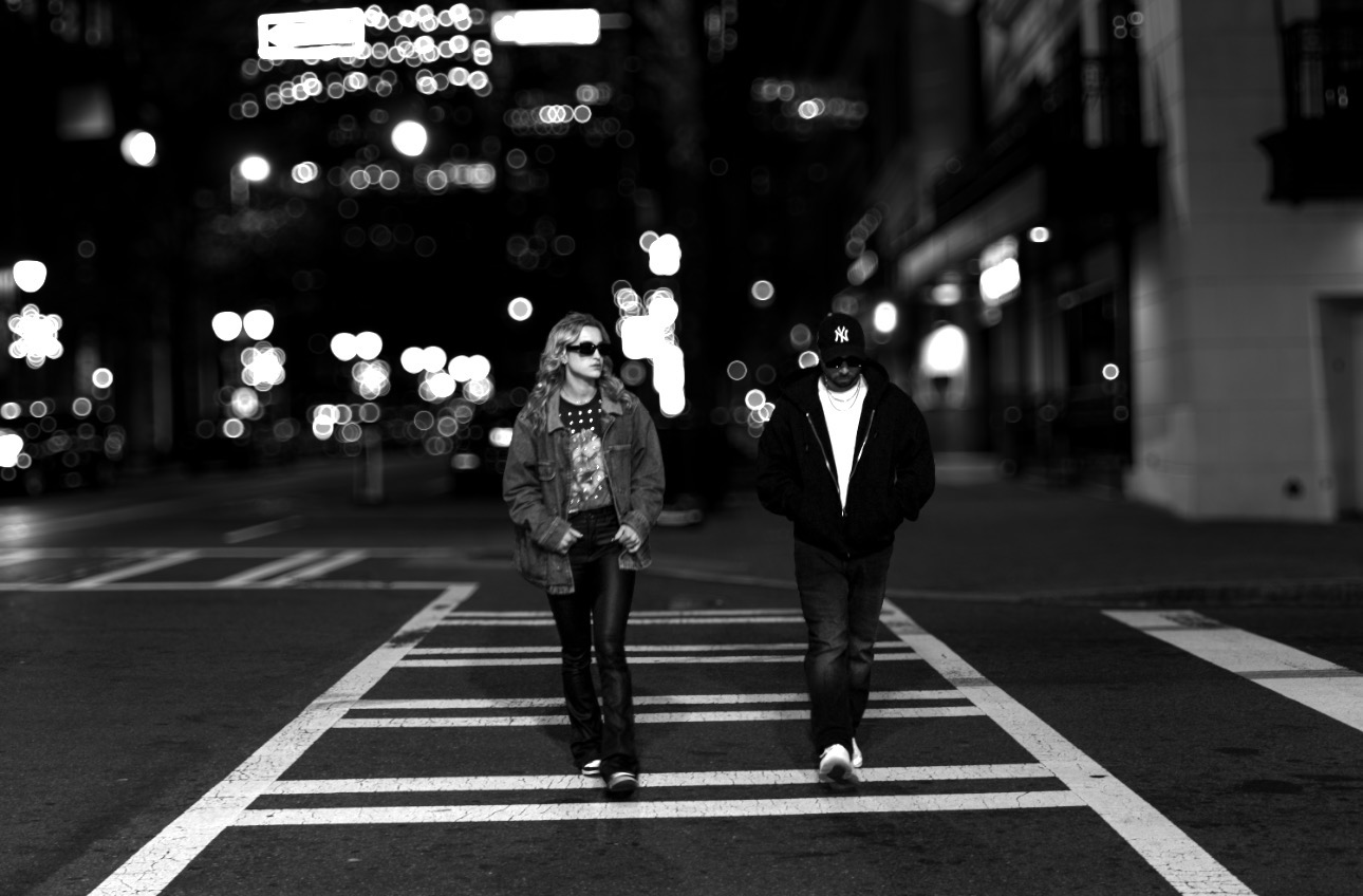 Two people walk across a city street at night with blurred city lights in the background.