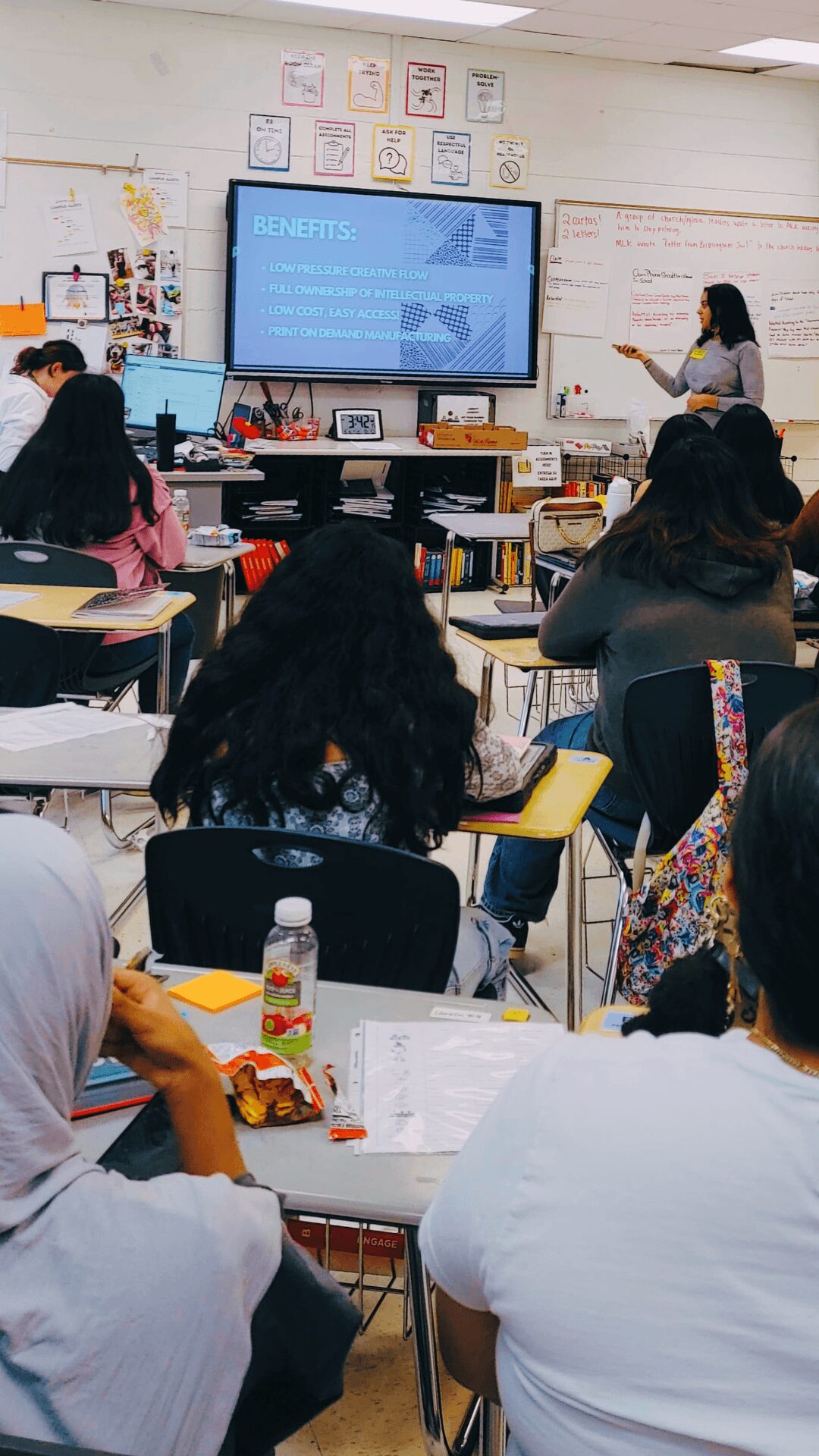 Classroom with students facing a teacher and a large screen displaying a presentation, whiteboard on the side, students taking notes.