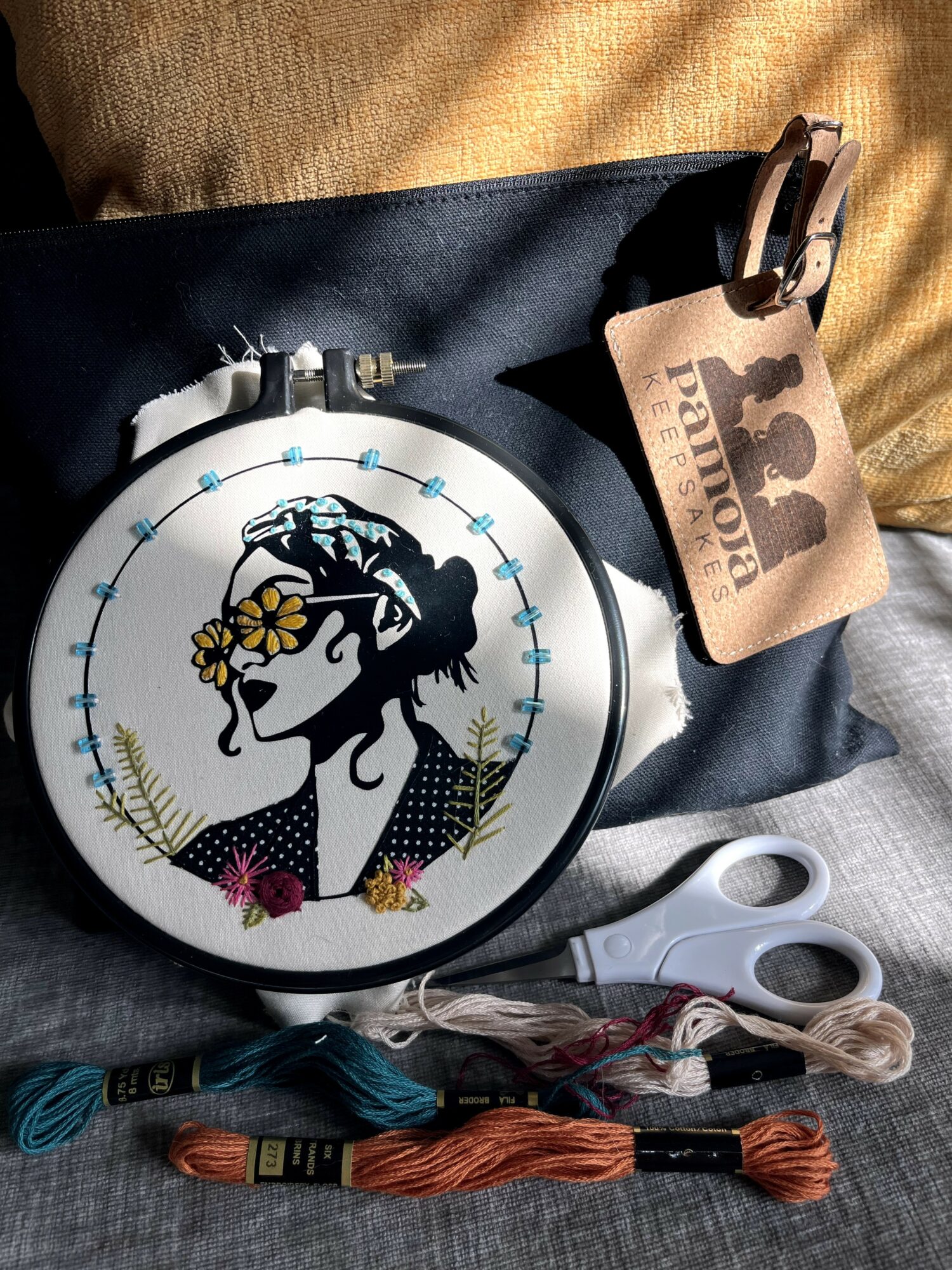 Embroidery hoop with a silhouette of a woman wearing glasses and flowers, scissors, thread, and a tag with a paw print, on fabric.