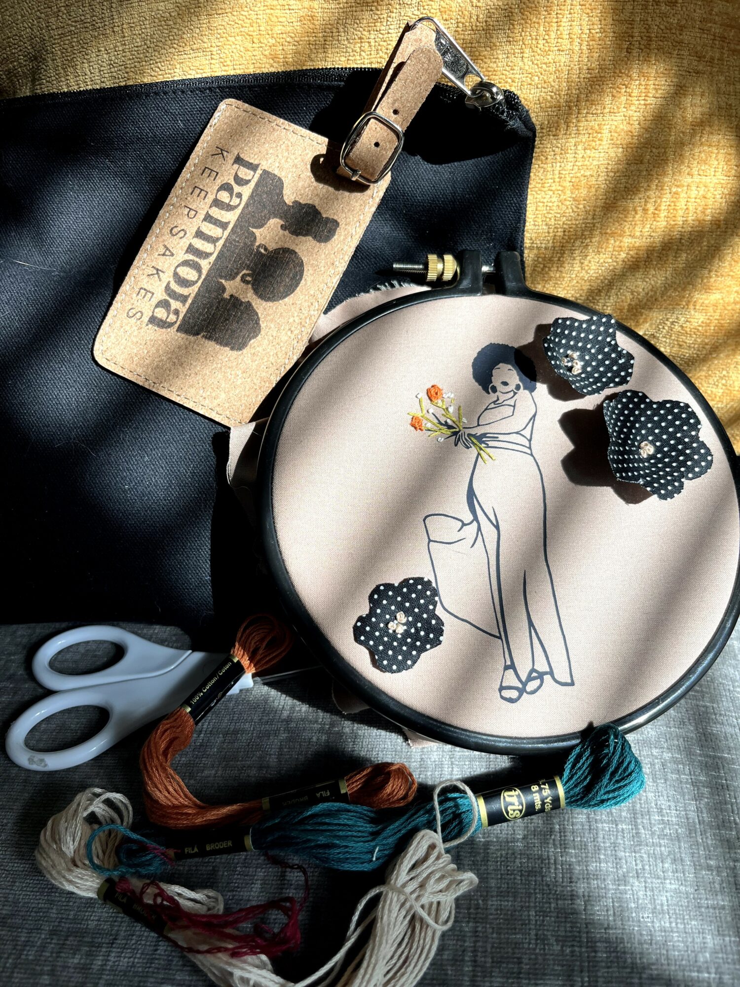 Embroidery hoop with a partially stitched design of a woman, surrounded by fabric pieces, scissors, and embroidery threads on fabric.