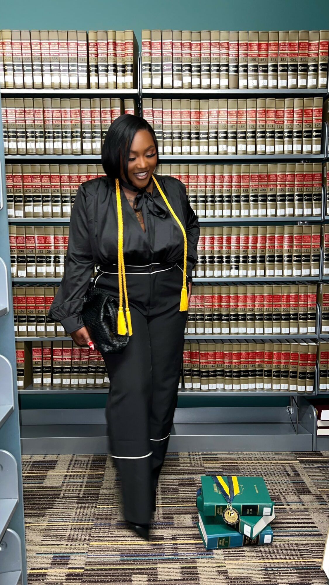 Woman standing in front of a bookshelf, smiling, with a small robot on the floor, wearing a black outfit and yellow measuring tape.