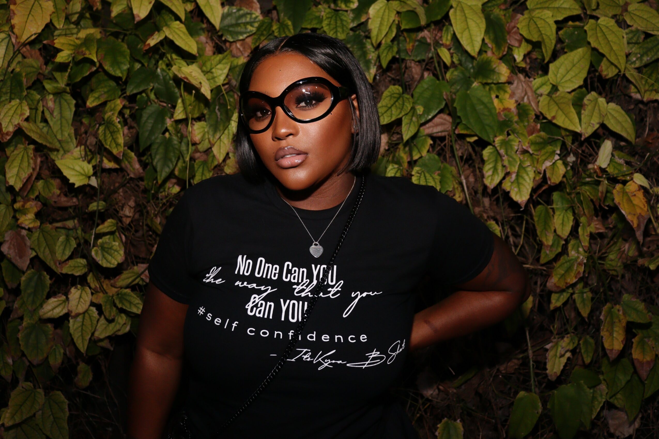 Woman with glasses and short hair poses in front of leafy background, wearing a black T-shirt with white text.