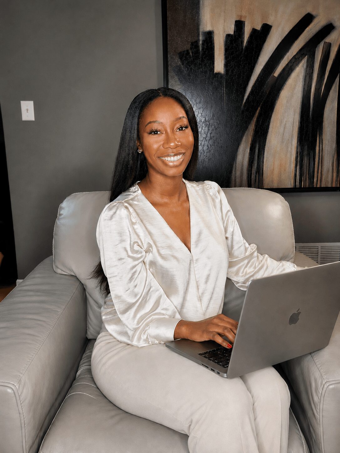 Smiling woman sitting on a sofa with a laptop, wearing a light-colored blouse, in a modern room with artwork on the wall.